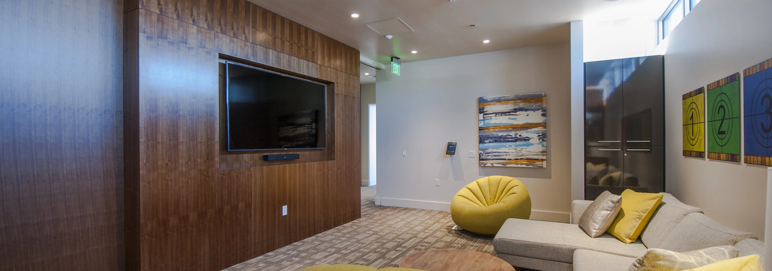 Media lounge at AMLI Waterfront apartments with flat-screen TV, wood accent wall, yellow chairs, and modern sectional sofa.