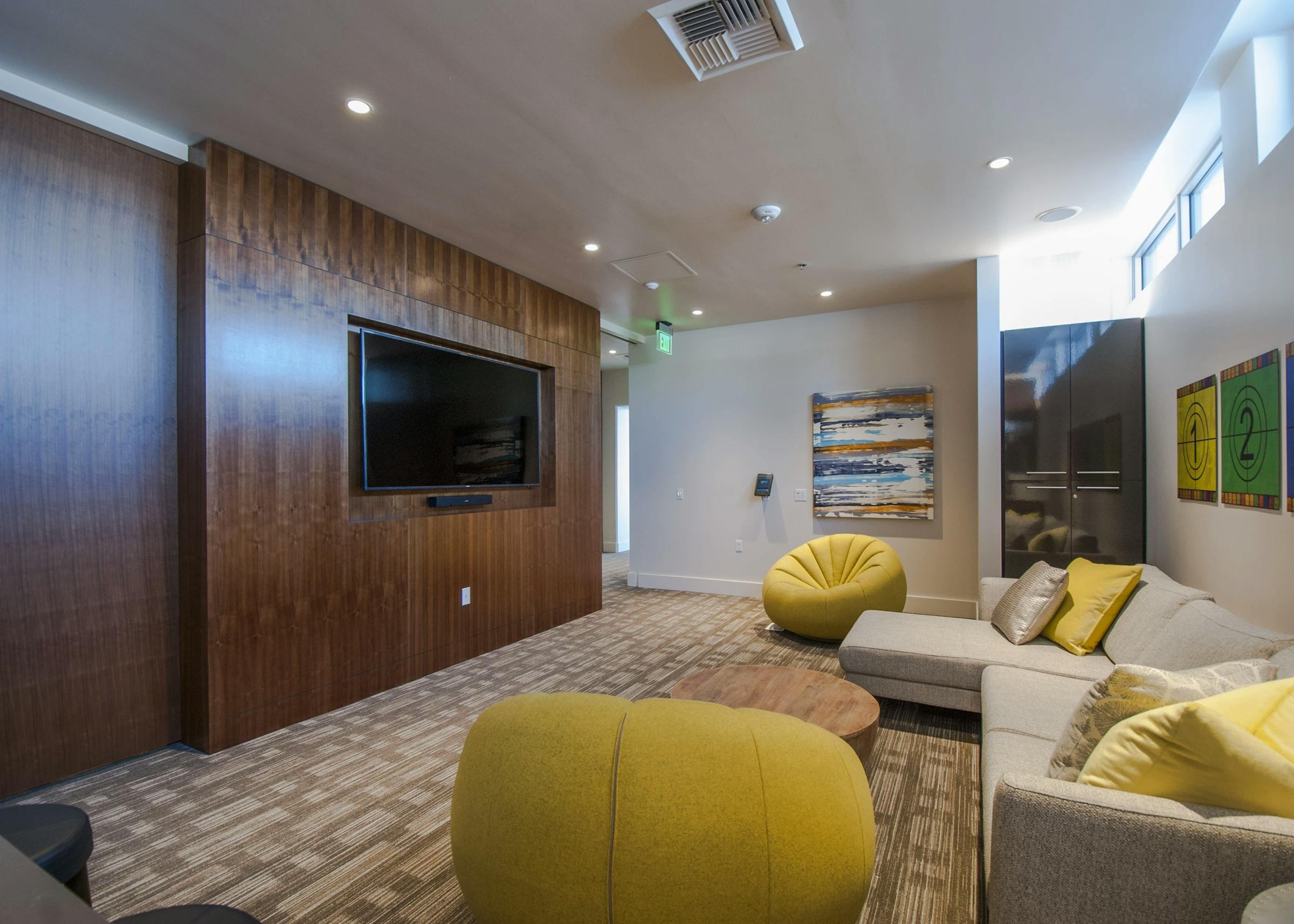Media lounge at AMLI Waterfront apartments with flat-screen TV, wood accent wall, yellow chairs, and modern sectional sofa.