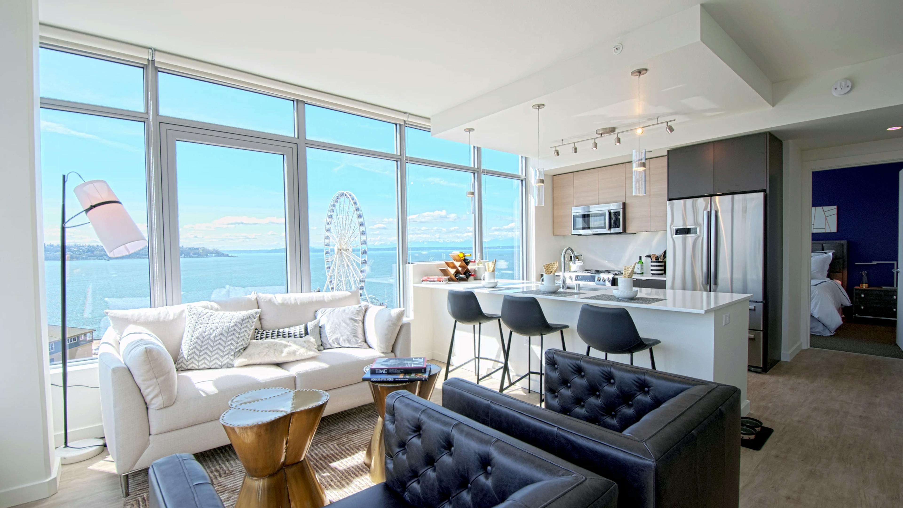 Open AMLI Waterfront apartment with black armchairs, white sofa, kitchen island, and ferris wheel view through wall of windows.