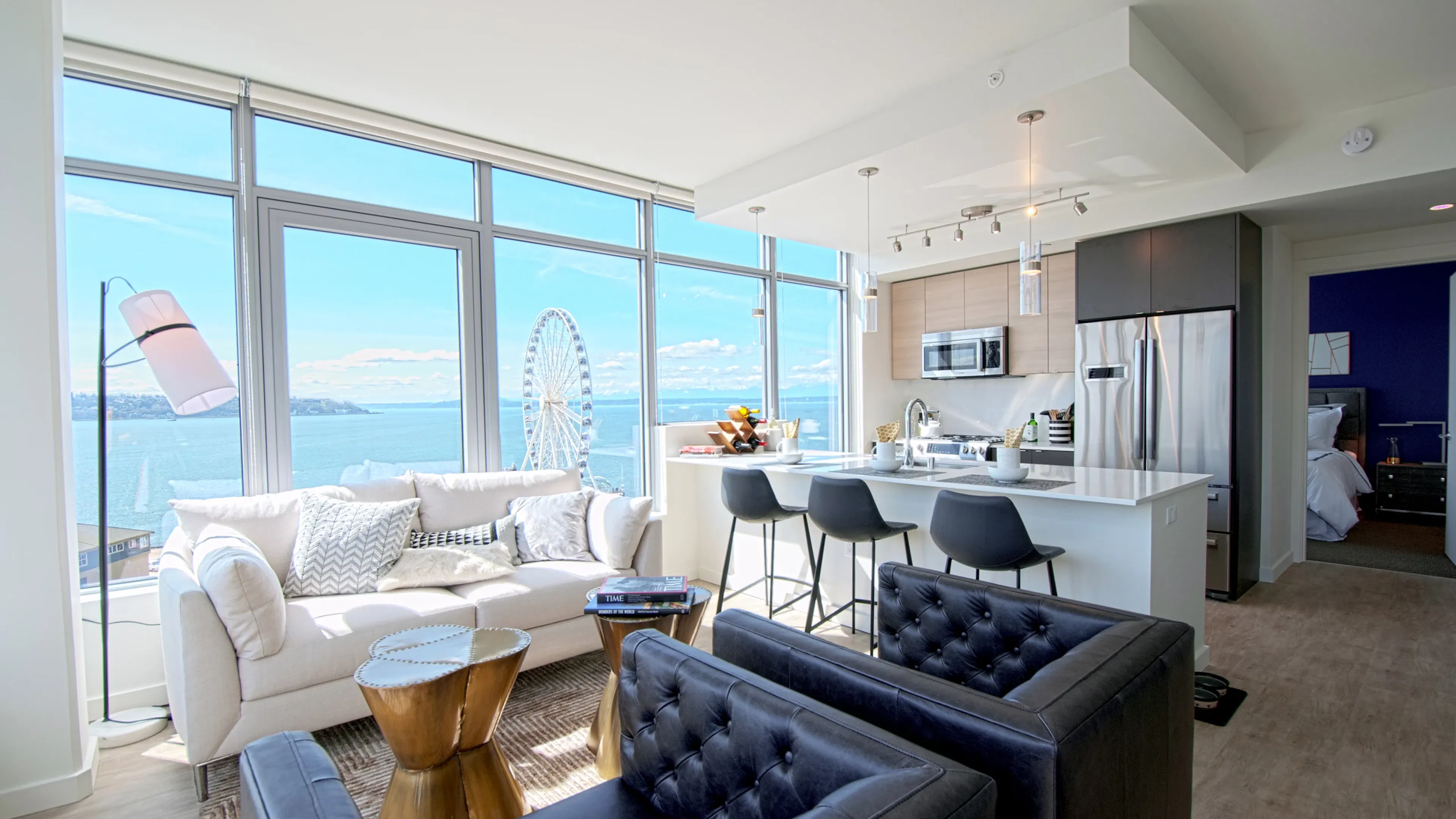 Open AMLI Waterfront apartment with black armchairs, white sofa, kitchen island, and ferris wheel view through wall of windows.