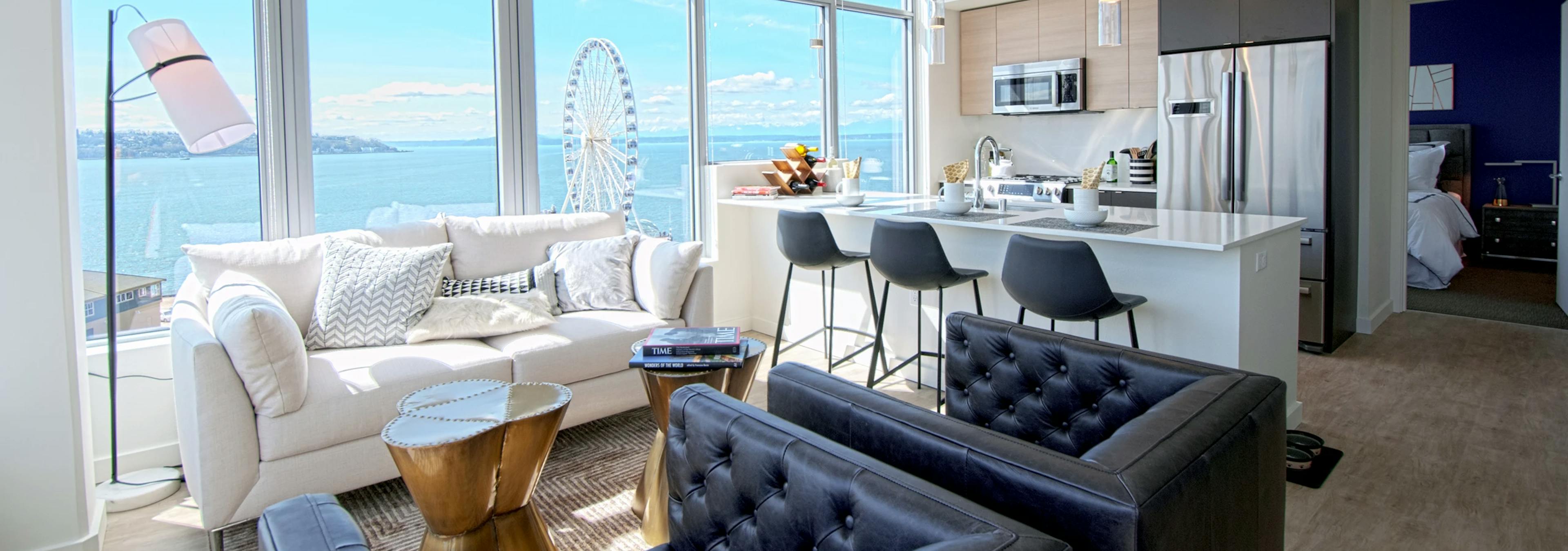 Open AMLI Waterfront apartment with black armchairs, white sofa, kitchen island, and ferris wheel view through wall of windows.