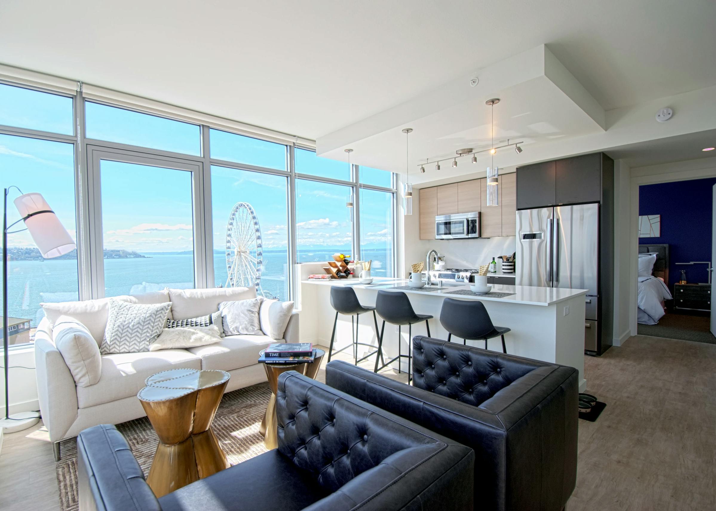 Open AMLI Waterfront apartment with black armchairs, white sofa, kitchen island, and ferris wheel view through wall of windows.