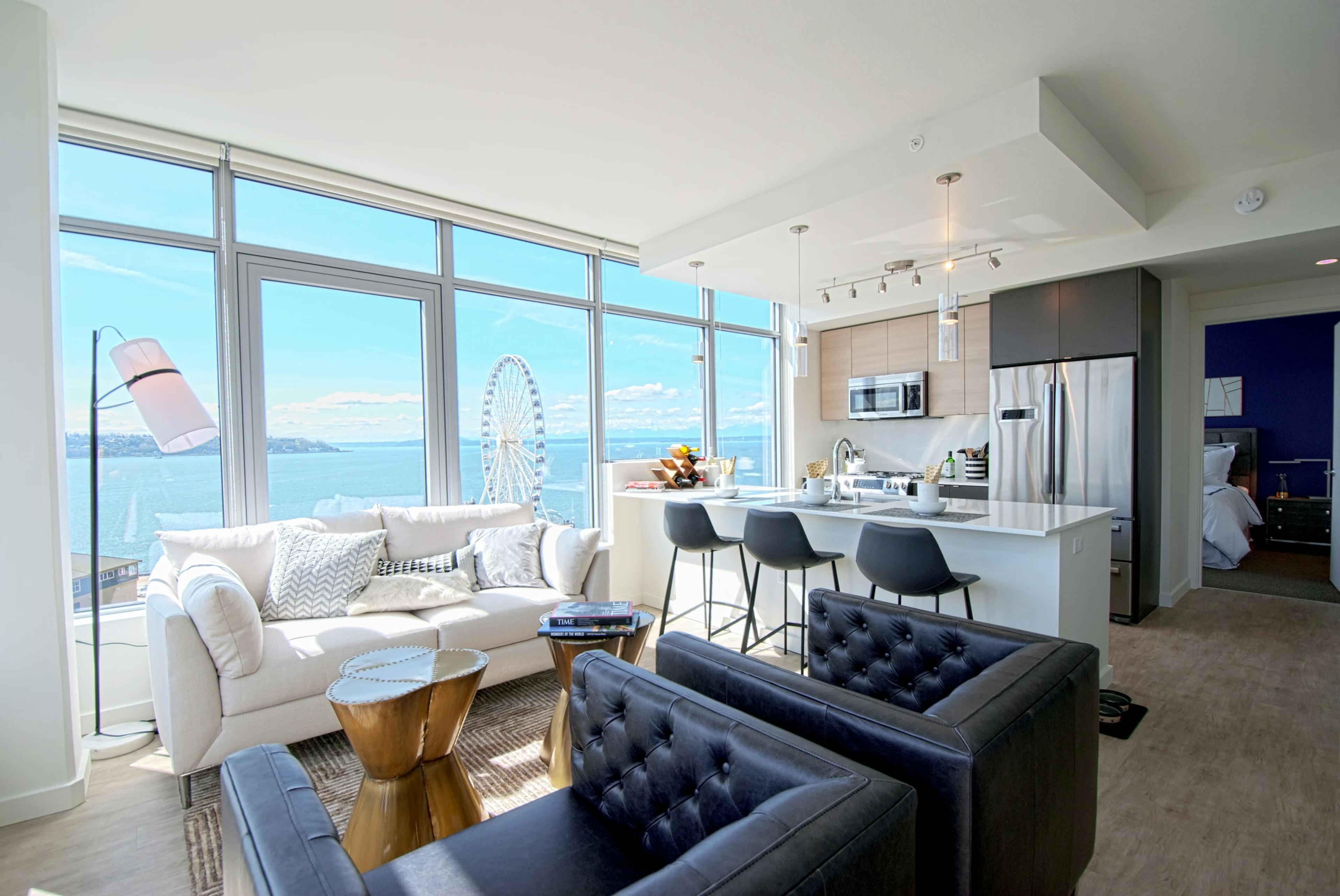 Open AMLI Waterfront apartment with black armchairs, white sofa, kitchen island, and ferris wheel view through wall of windows.
