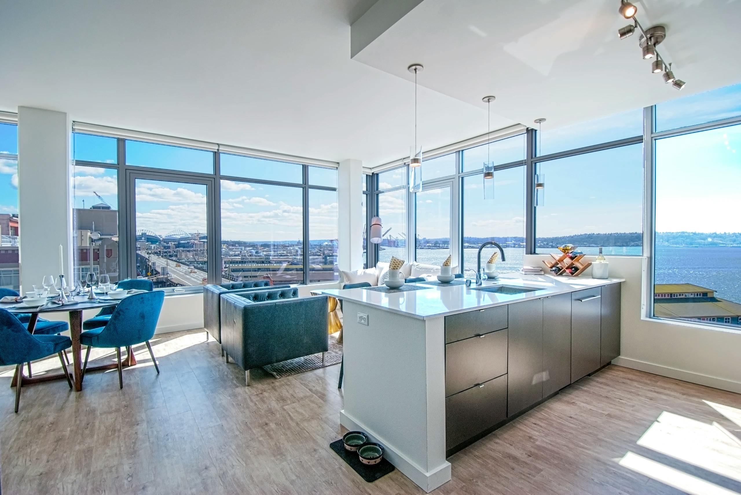 Open living area at AMLI Waterfront apartments with kitchen island, large corner windows, blue dining chairs, and city and ocean views.