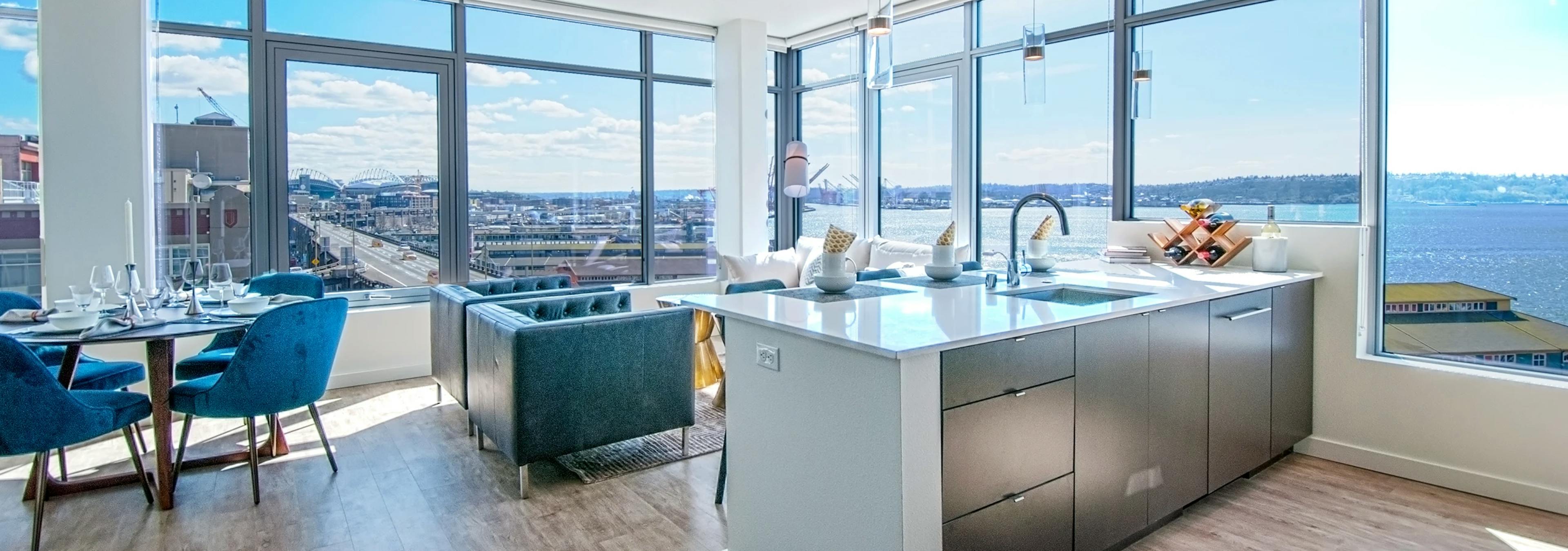 Open living area at AMLI Waterfront apartments with kitchen island, large corner windows, blue dining chairs, and city and ocean views.