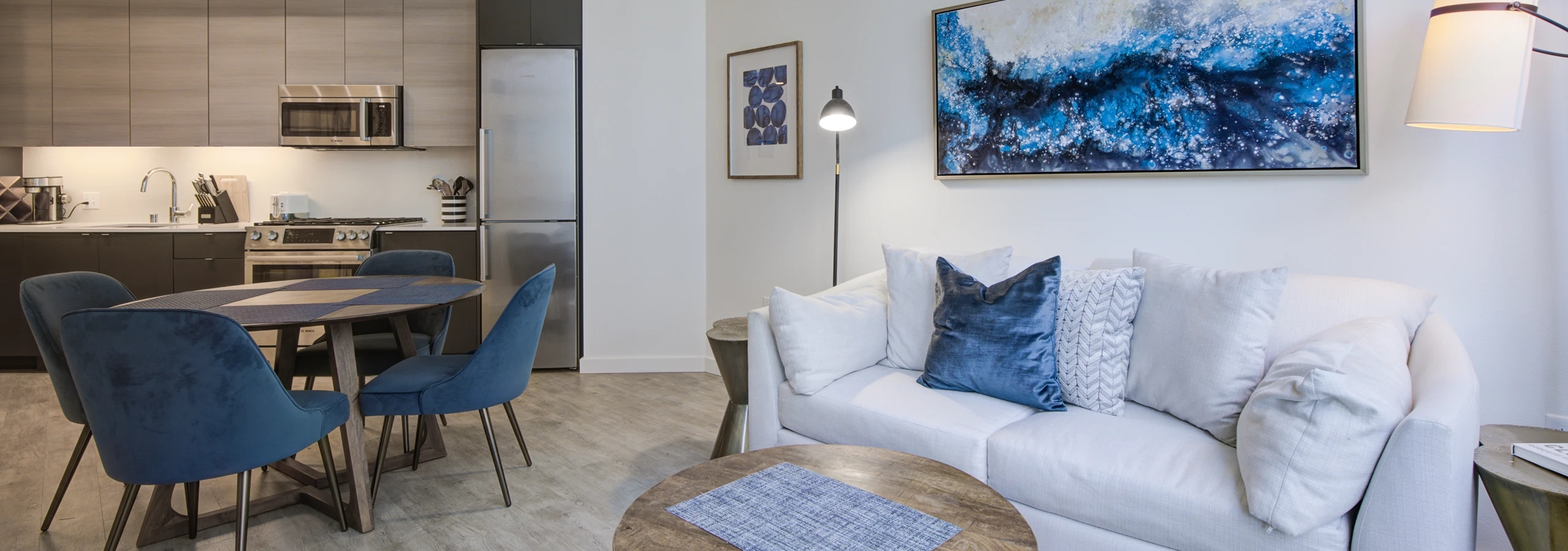 Living room and kitchen at AMLI Waterfront apartments with white sofa, blue chairs, round wood table and light wood cabinets.