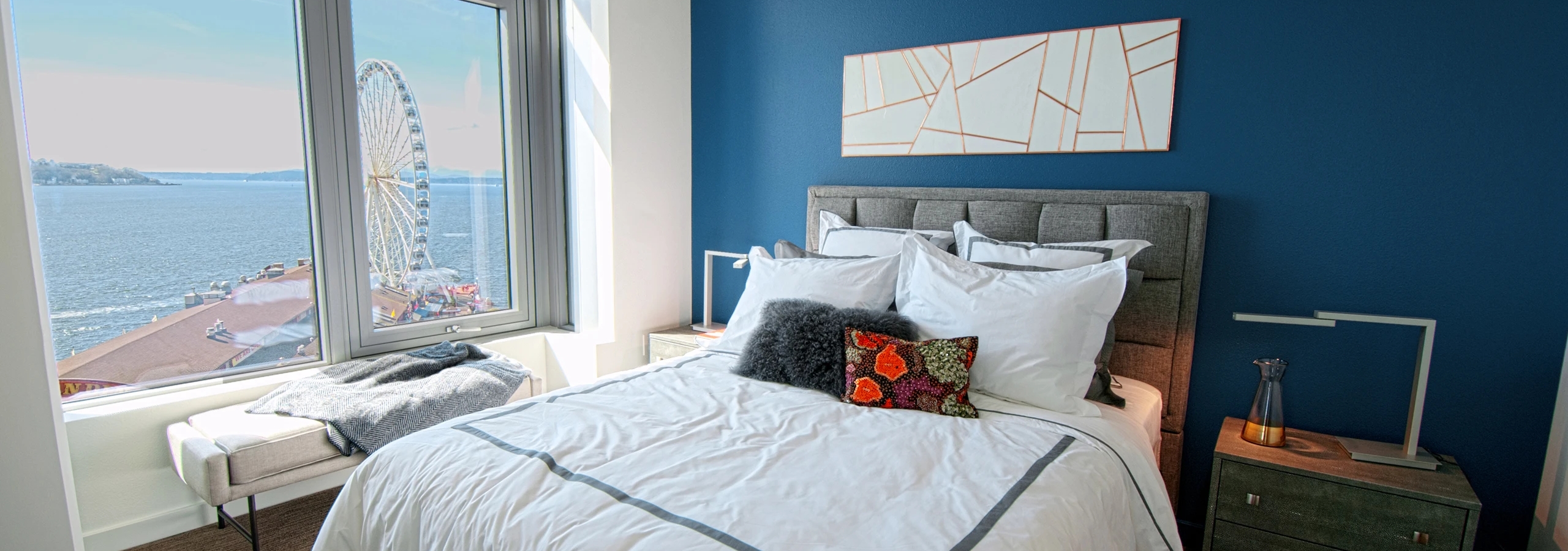 AMLI Waterfront apartment bedroom with ocean and ferris wheel view, blue accent wall, gray bed frame, and white bedding with pillows.