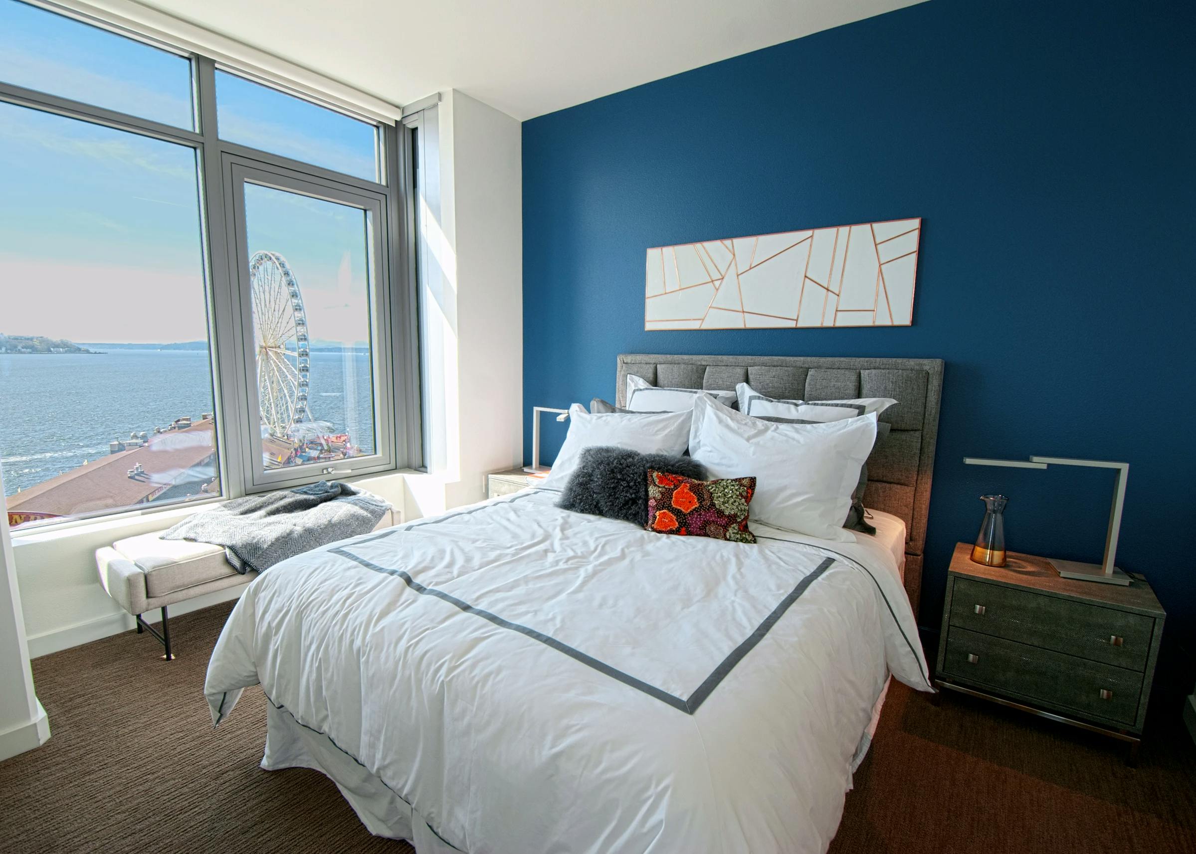 AMLI Waterfront apartment bedroom with ocean and ferris wheel view, blue accent wall, gray bed frame, and white bedding with pillows.