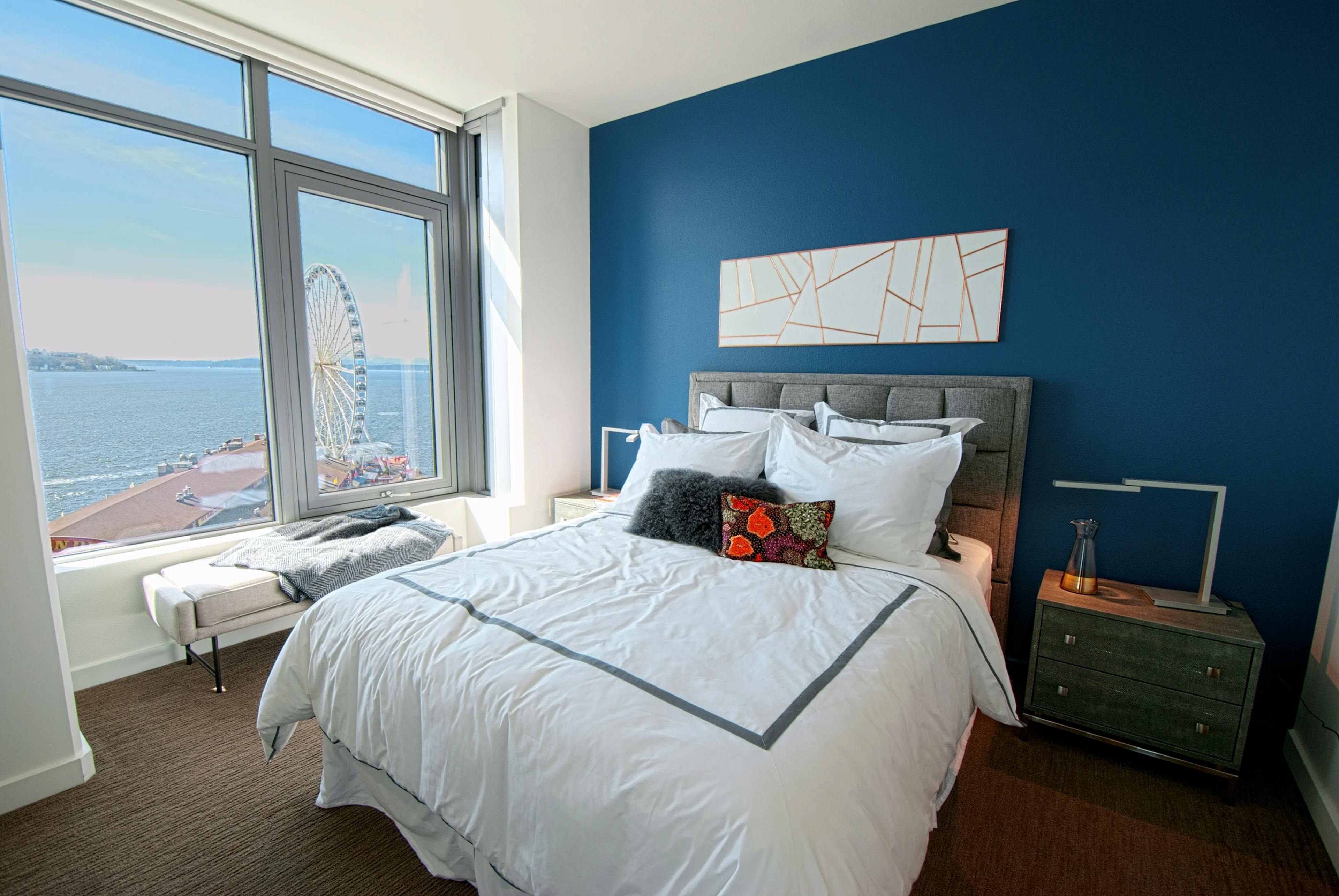 AMLI Waterfront apartment bedroom with ocean and ferris wheel view, blue accent wall, gray bed frame, and white bedding with pillows.