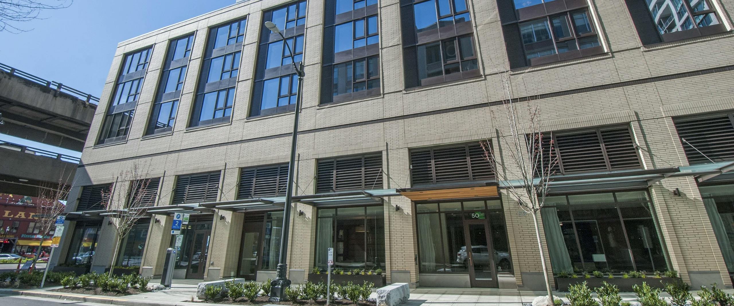 Daytime exterior of AMLI Waterfront apartment building entrance with brick and glass facade and trees.