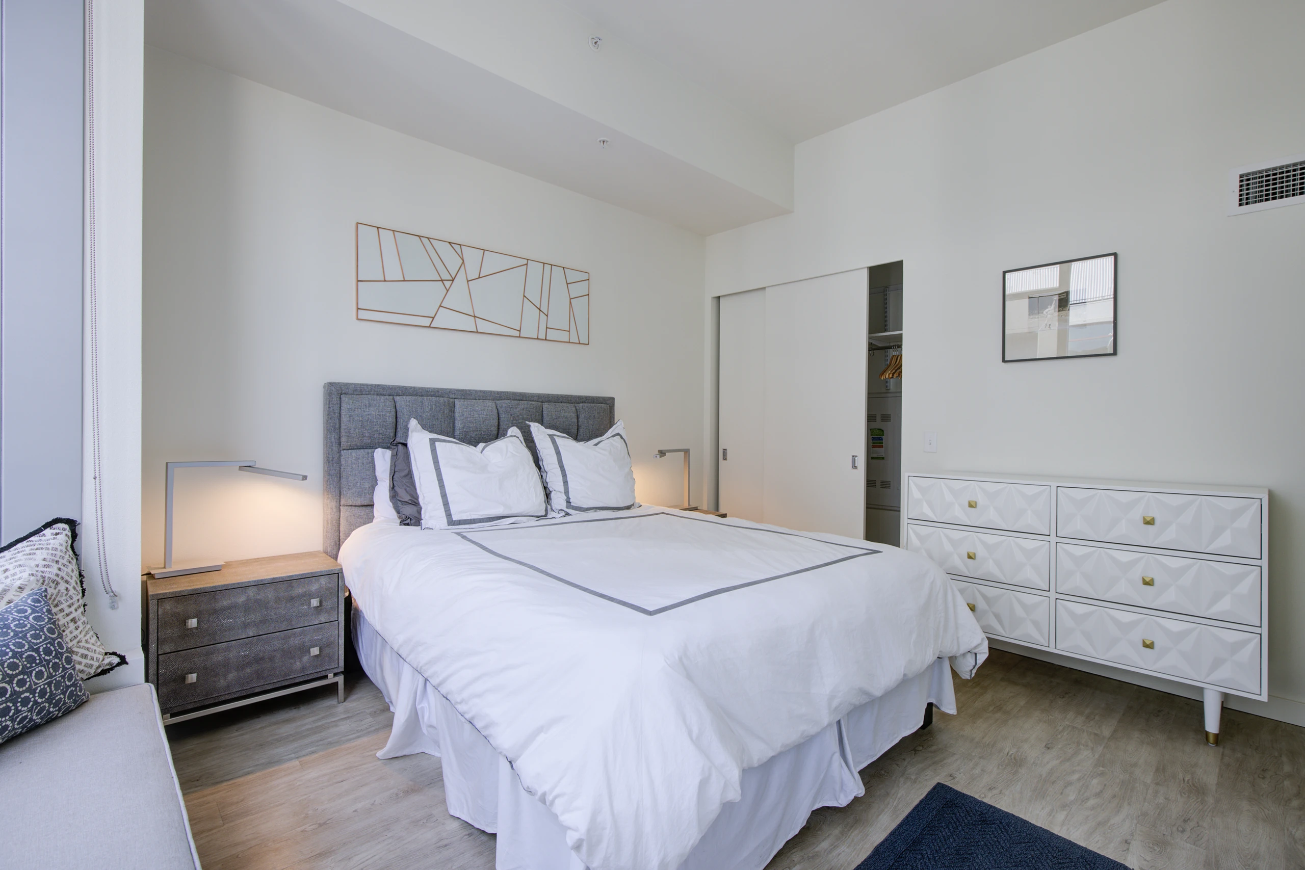 Interior view of AMLI Waterfront bedroom with gray headboard, white bedding, two nightstands, and white dresser with gold hardware.