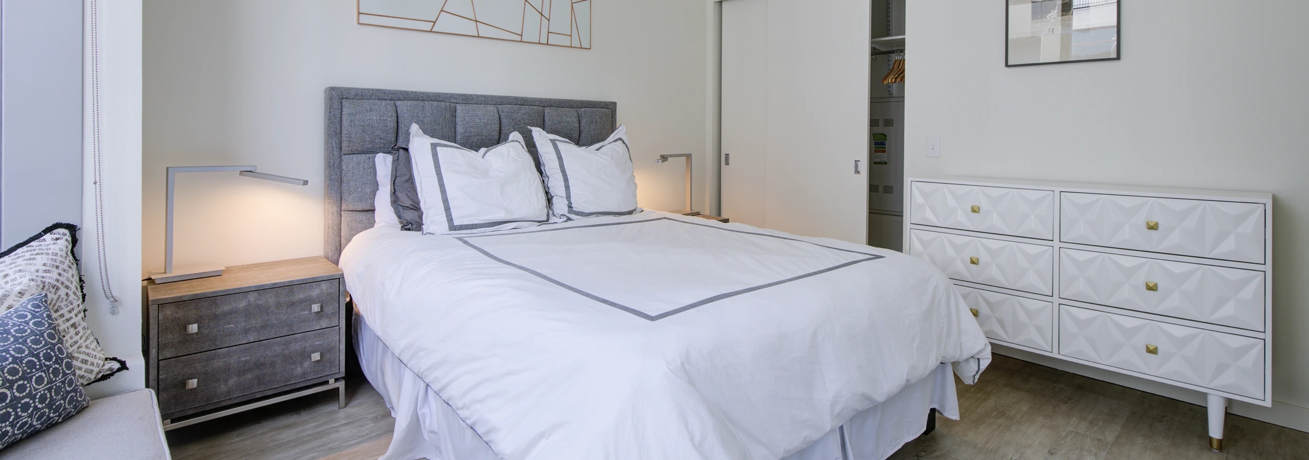 Interior view of AMLI Waterfront bedroom with gray headboard, white bedding, two nightstands, and white dresser with gold hardware.