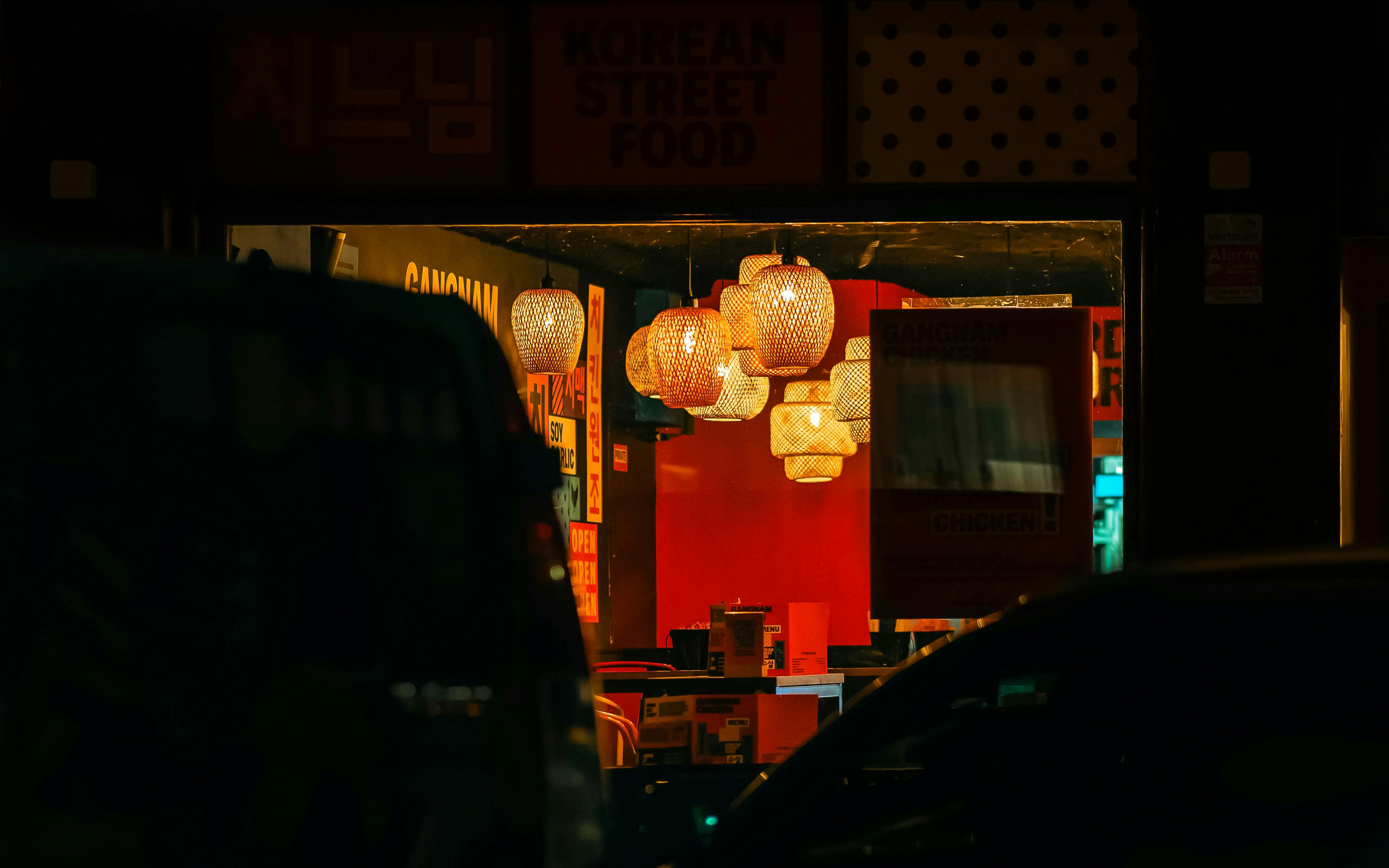 A dimly lit exterior shot of a restaurant at night, with hanging wicker-like lanterns casting a warm glow. The silhouettes of cars are in the foreground.