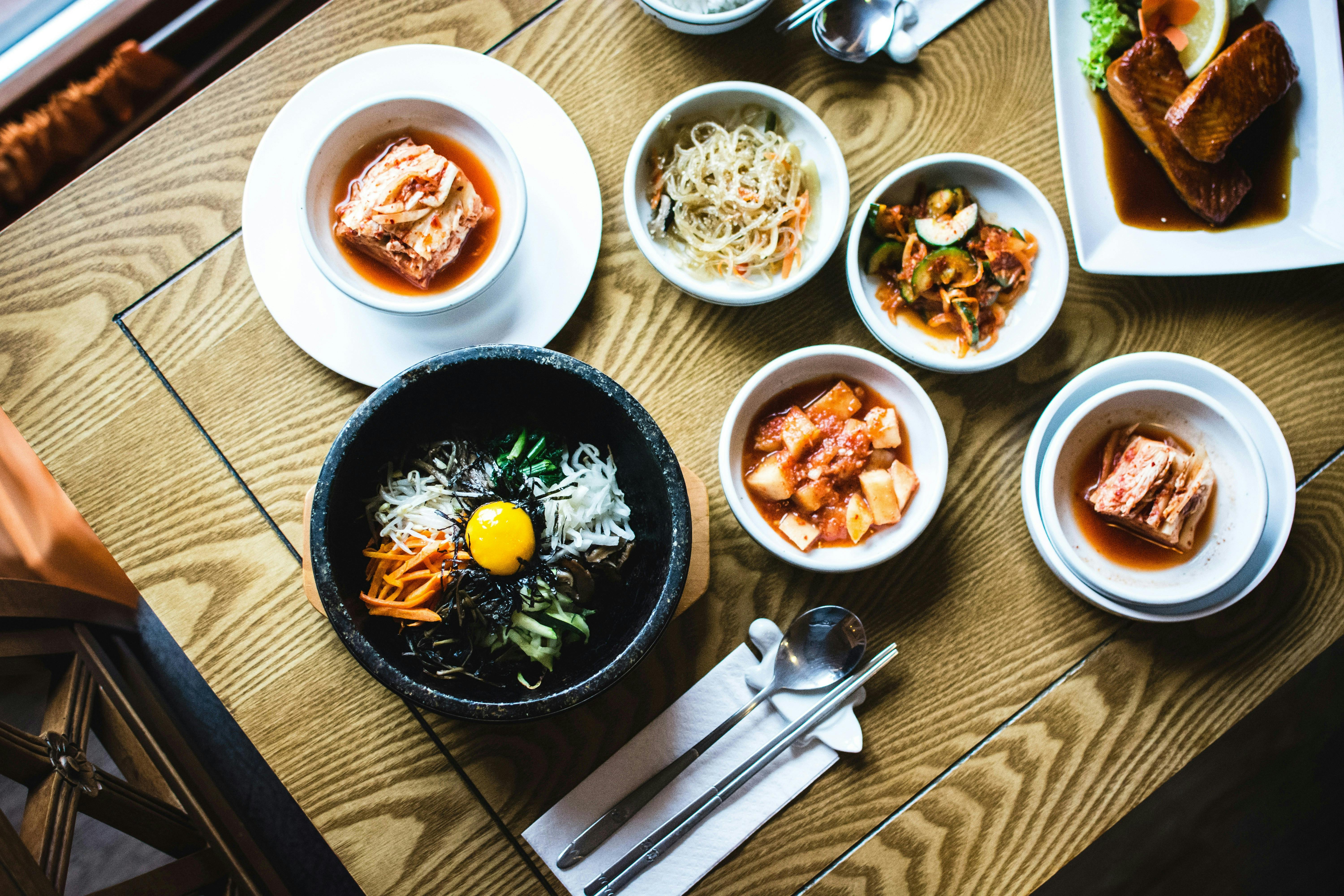 An overhead shot of a table with several small bowls of Korean food. A large black bowl in the center contains bibimbap with a raw egg yolk on top. Other side dishes (banchan) include kimchi and various vegetables.