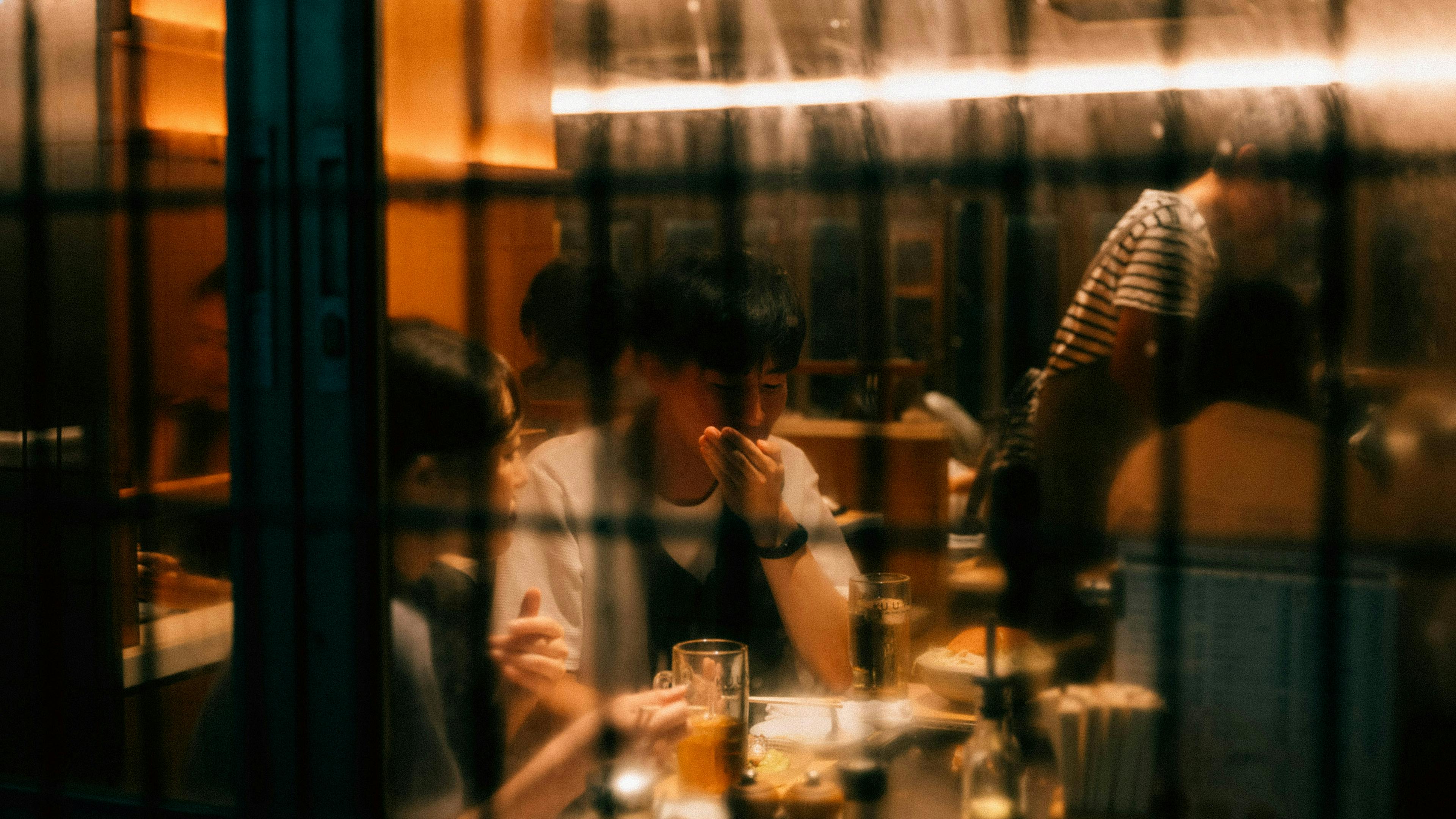 A group of people dining at a restaurant, viewed through a window with a metallic grate. The scene is warmly lit, with a man in the center holding his hand to his chin.
