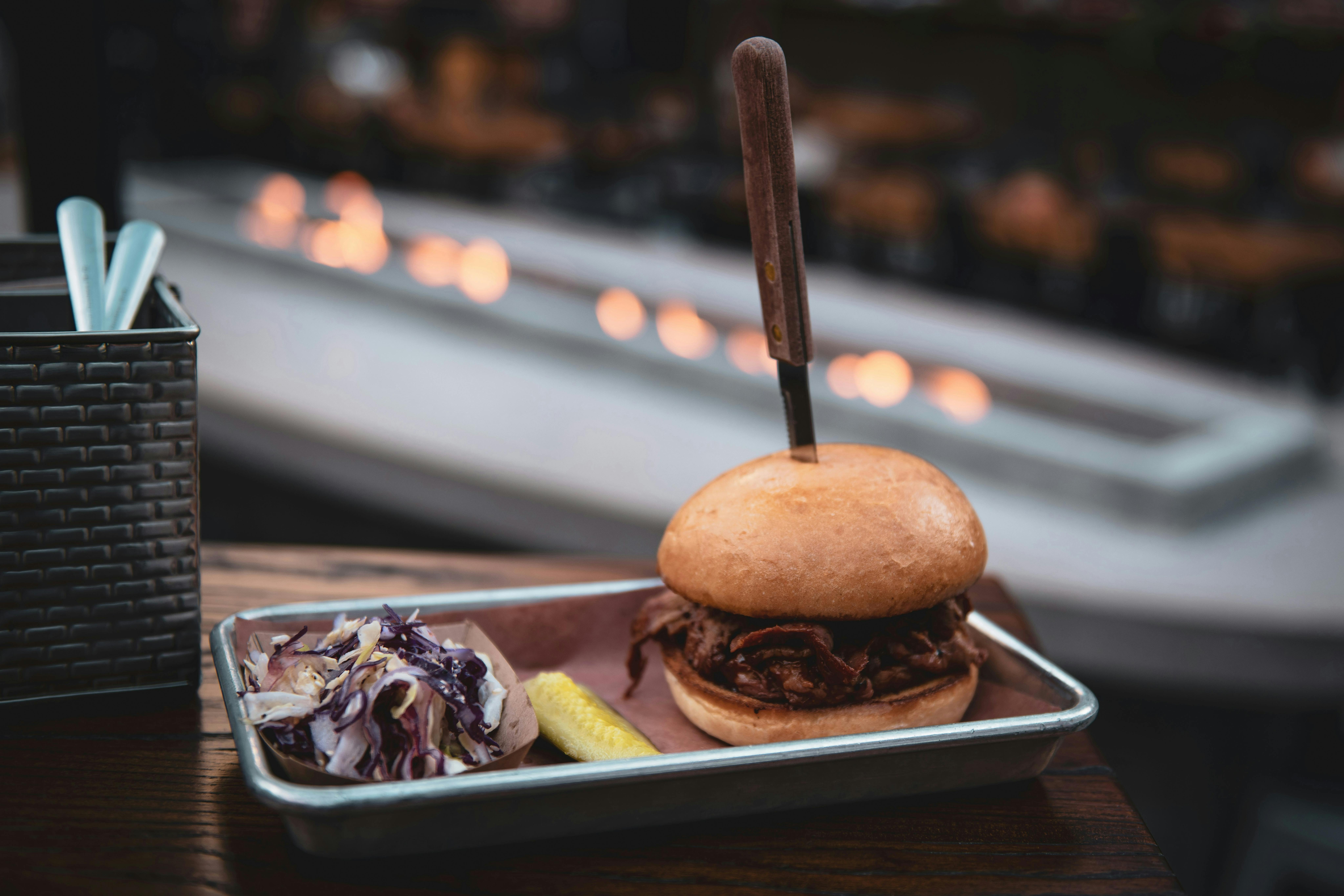 A gourmet pulled pork sandwich on a brioche bun, held together by a wooden knife, served on a metal tray with a side of coleslaw and a pickle. The background is a blurry outdoor patio with warm lights.