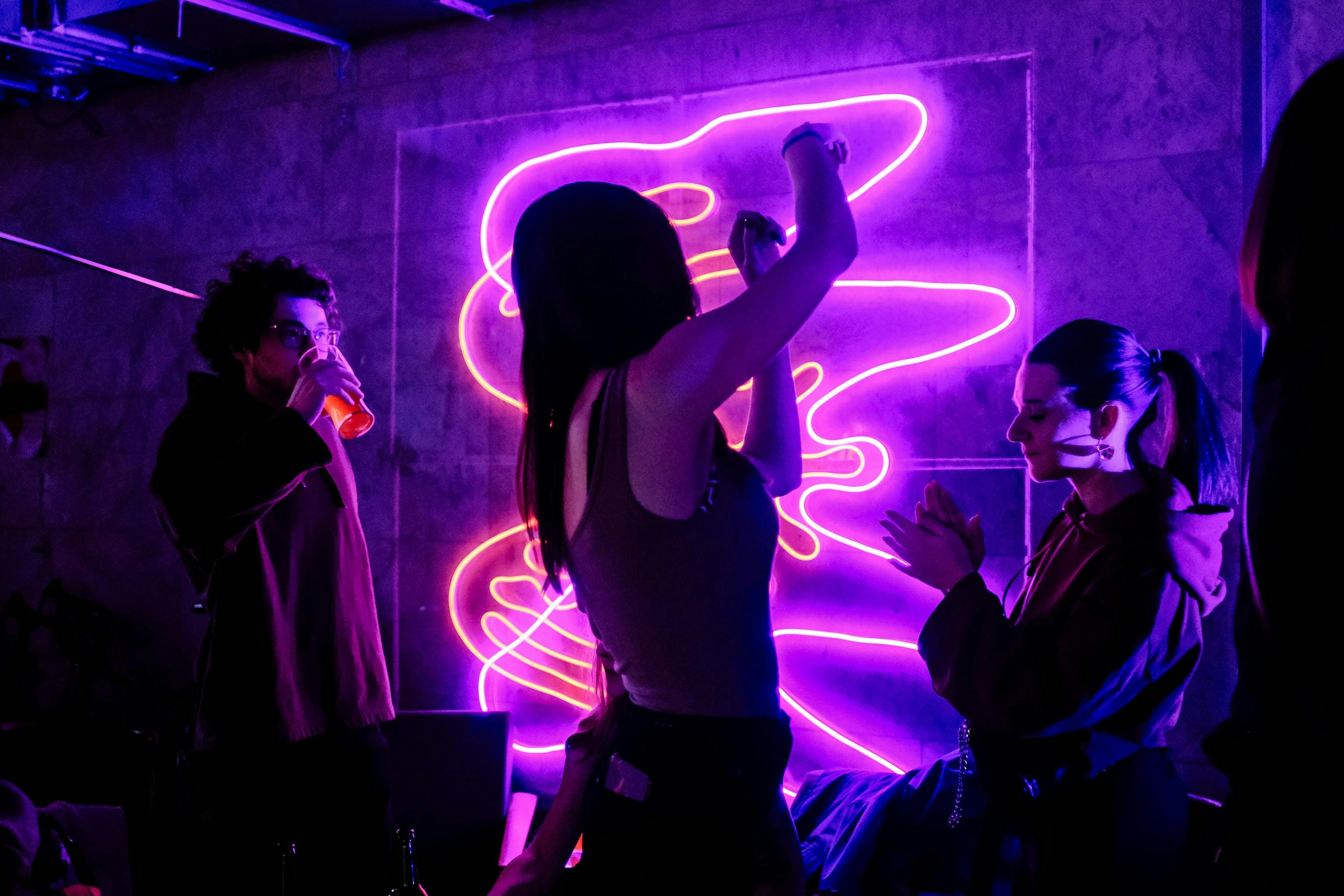 A group of people at a party or club, illuminated by a bright, magenta-colored neon light sculpture on the wall. A woman in the center is dancing with her arms raised.