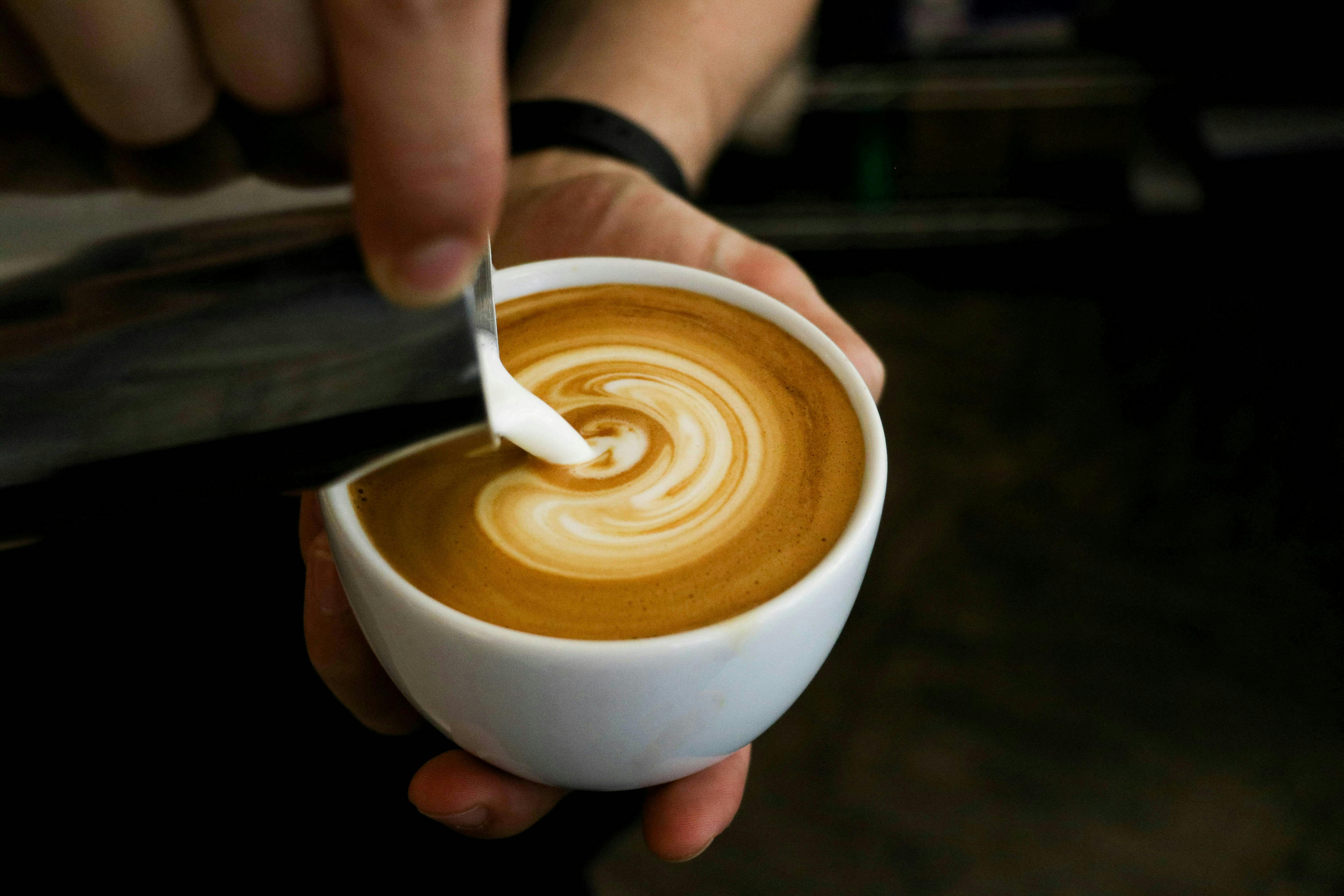 A close-up shot of a person's hands pouring steamed milk into a cup of coffee, creating latte art in a spiral pattern