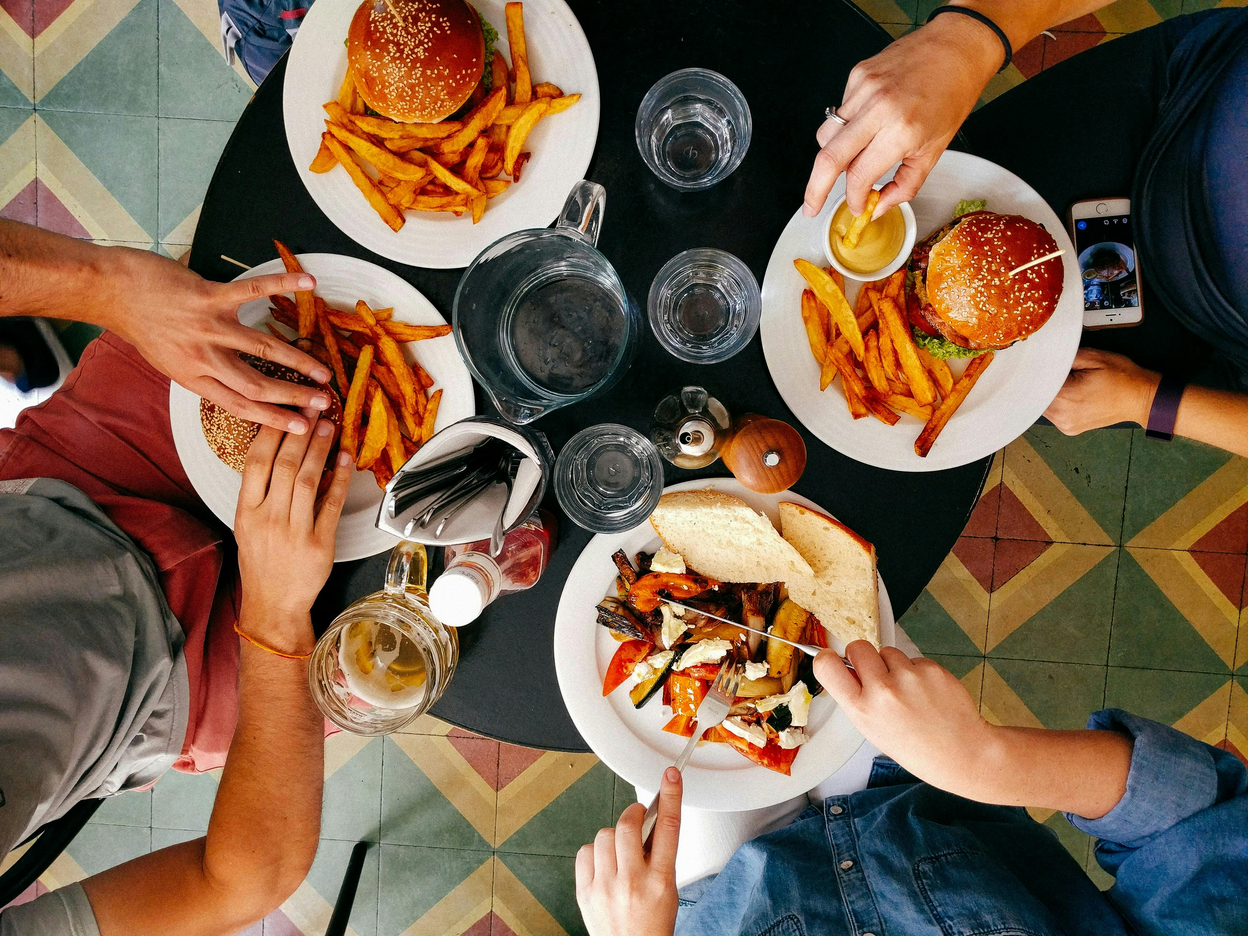 An overhead shot of a round table where four people are eating. The table holds two plates with burgers and fries, a plate with grilled vegetables and bread, and a pitcher of water with several glasses. Hands are visible reaching for food.