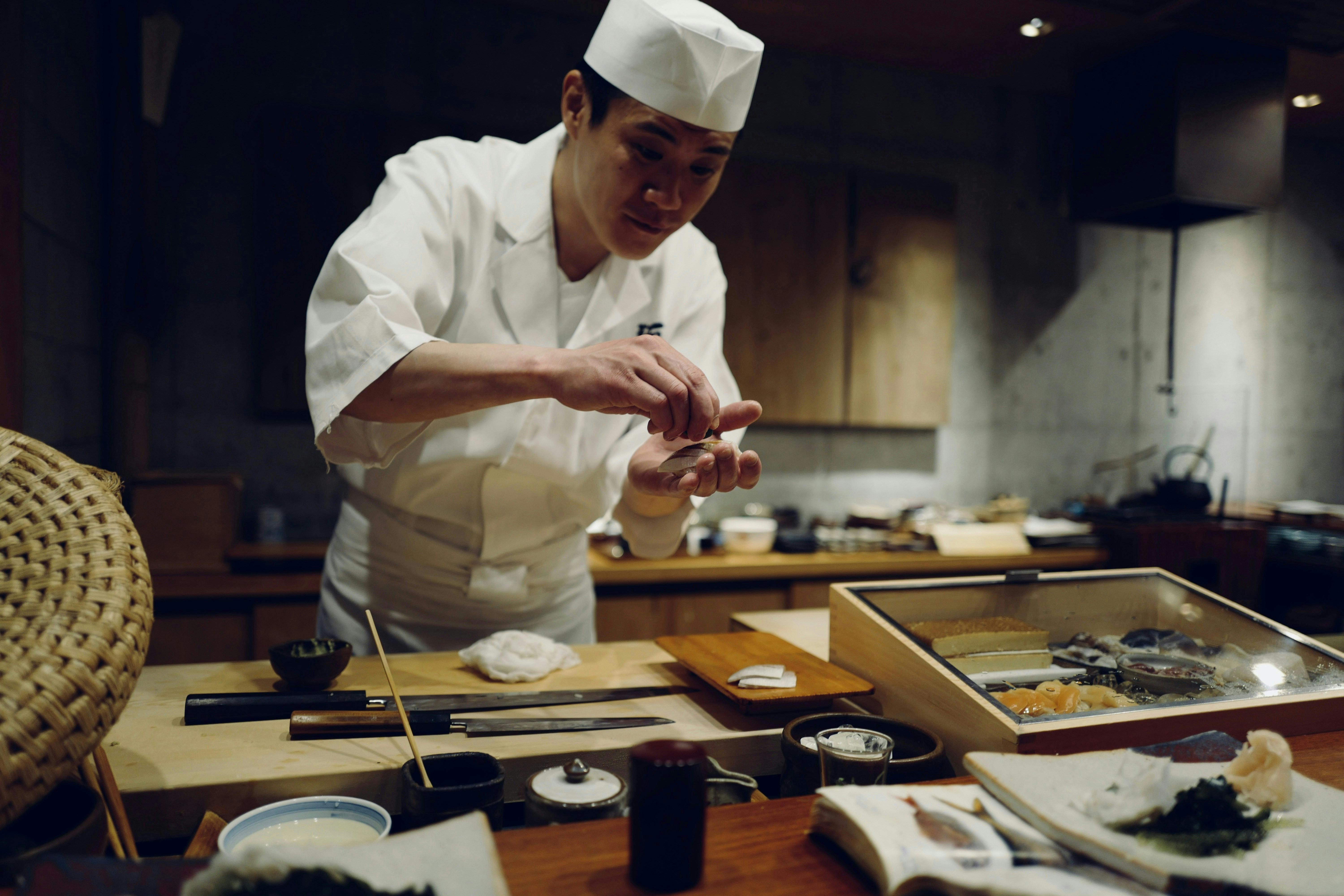 A Japanese sushi chef in a white uniform and hat is meticulously preparing sushi behind a wooden counter. His hands are delicately shaping a piece of fish or rice.