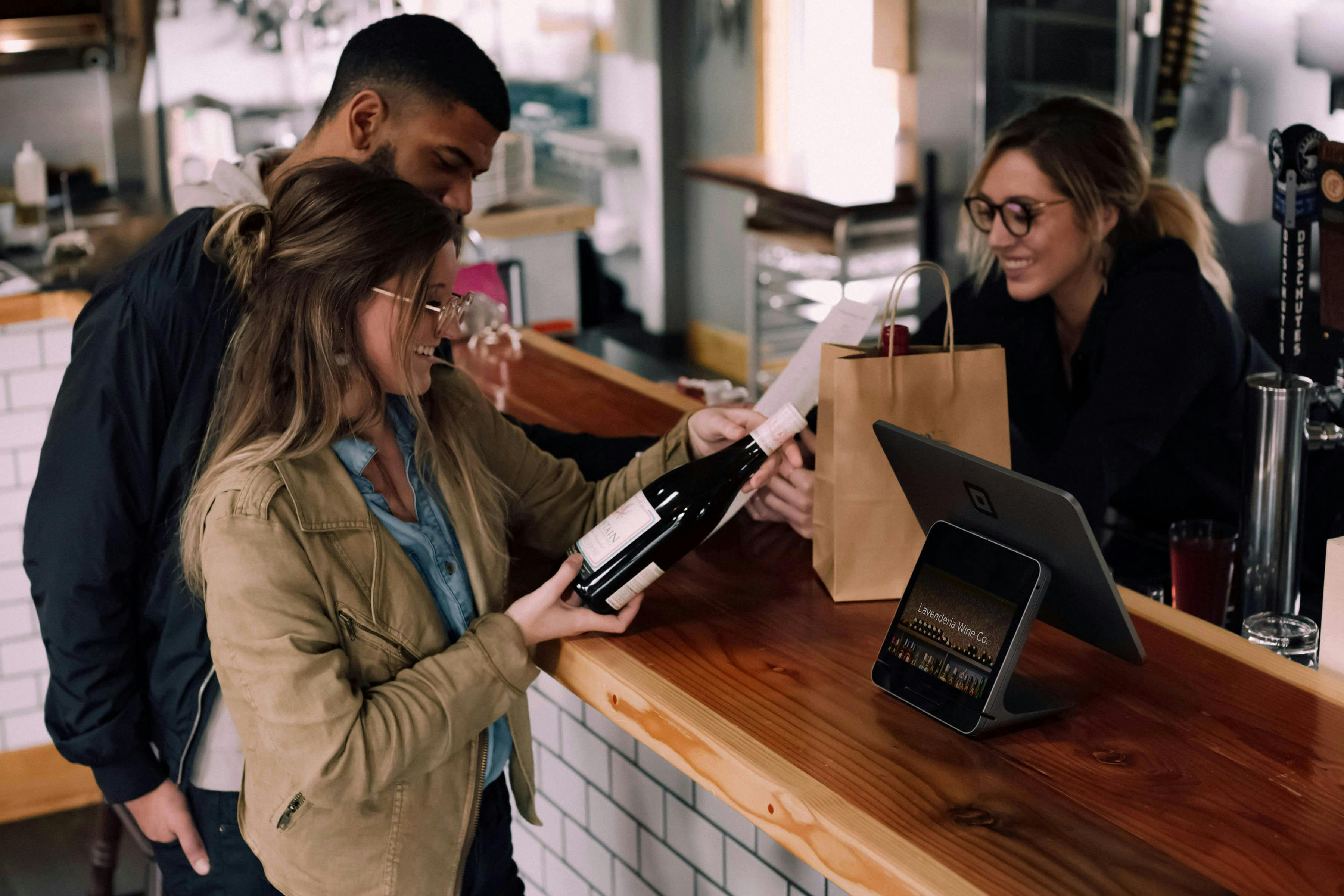 A customer at a bar counter is holding a bottle of wine and talking to a friendly bartender, with another customer standing beside them. A modern payment terminal is on the counter.