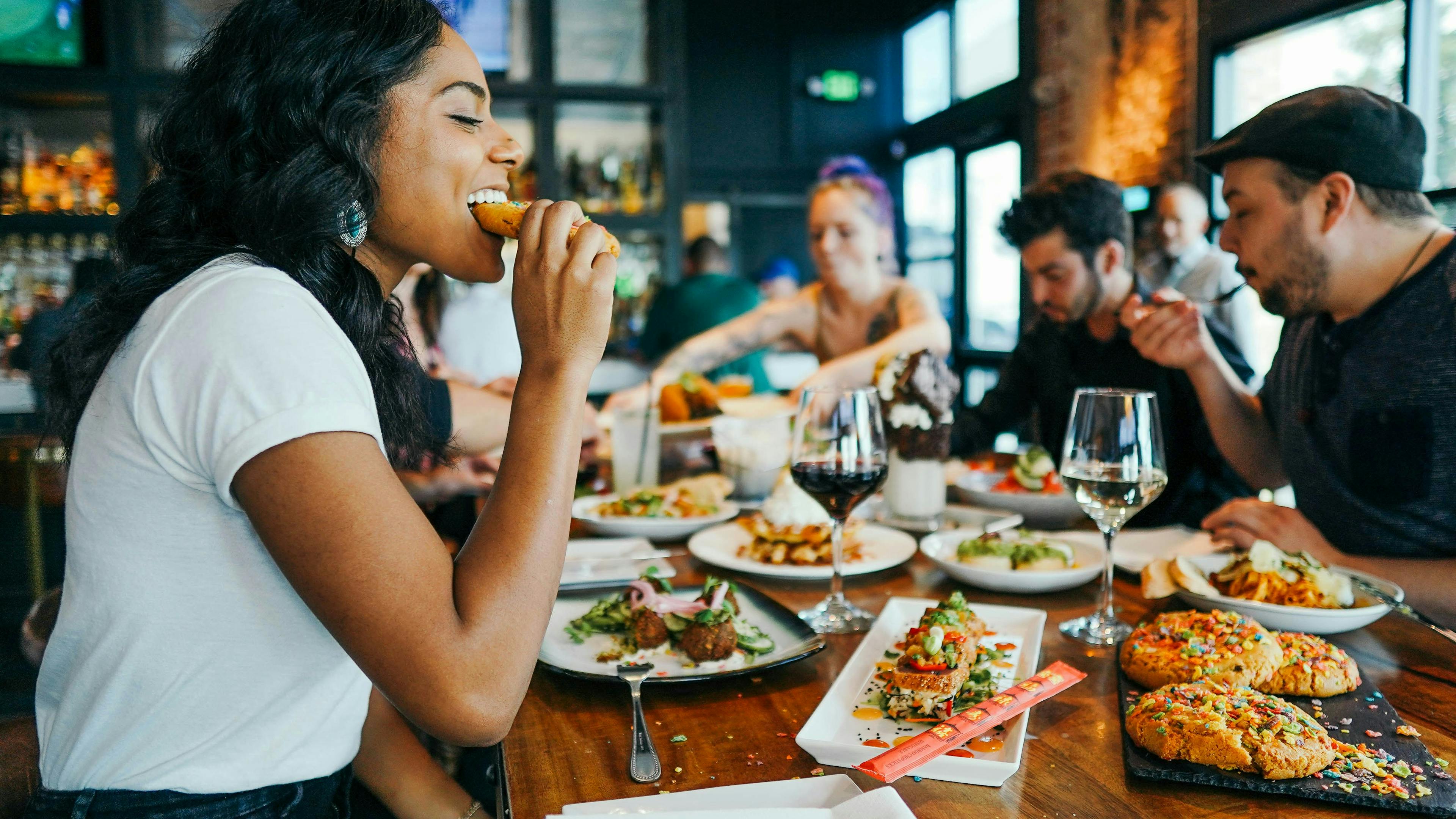 A group of friends sharing a meal at a restaurant. A woman in a white t-shirt on the left is taking a bite of food. The table is filled with plates of food, drinks, and a few people are talking and laughing in the background.