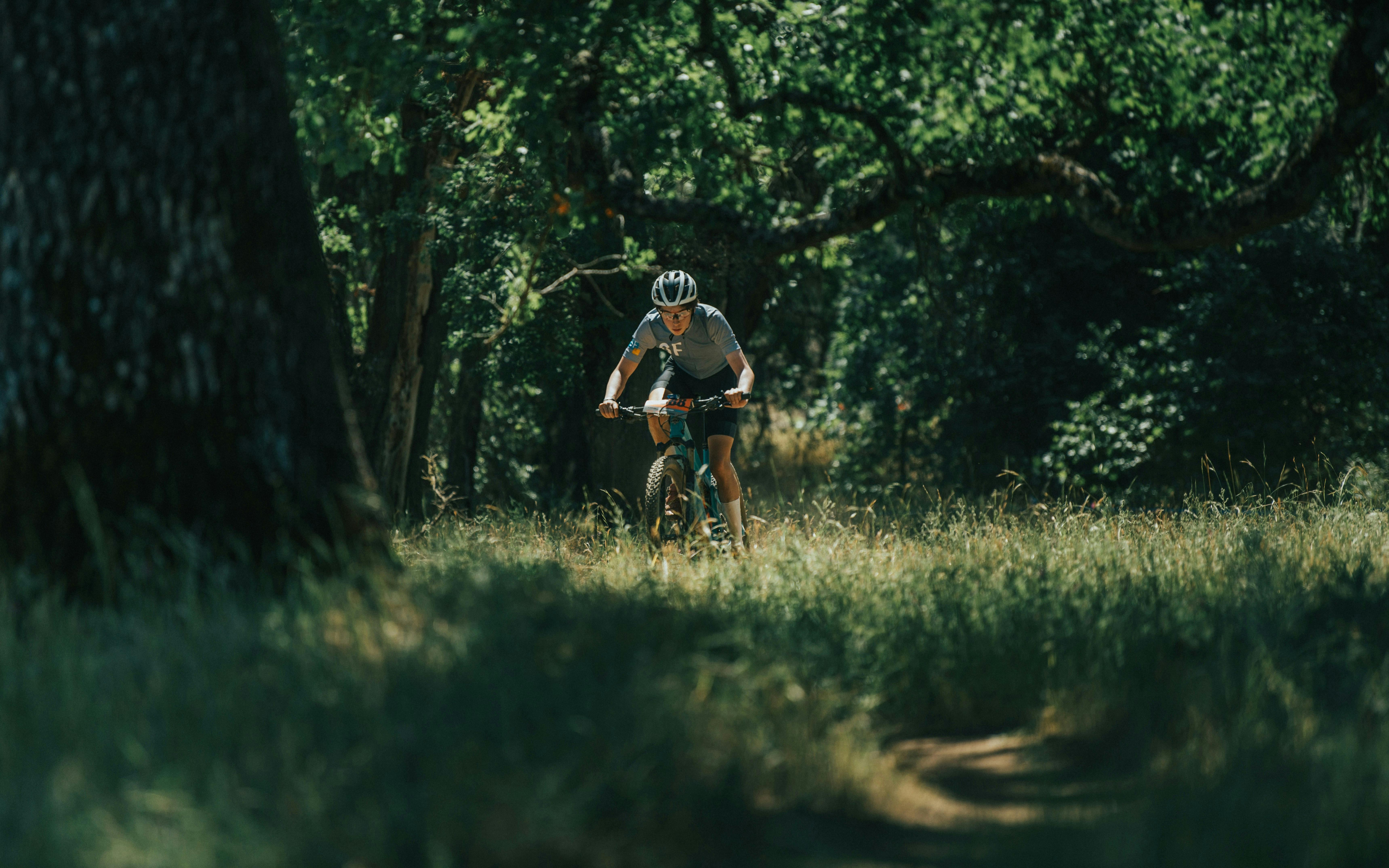 A person on a mountain bike is riding on a dirt path through a grassy, sun-dappled forest. The biker is wearing a helmet and a light-colored shirt.