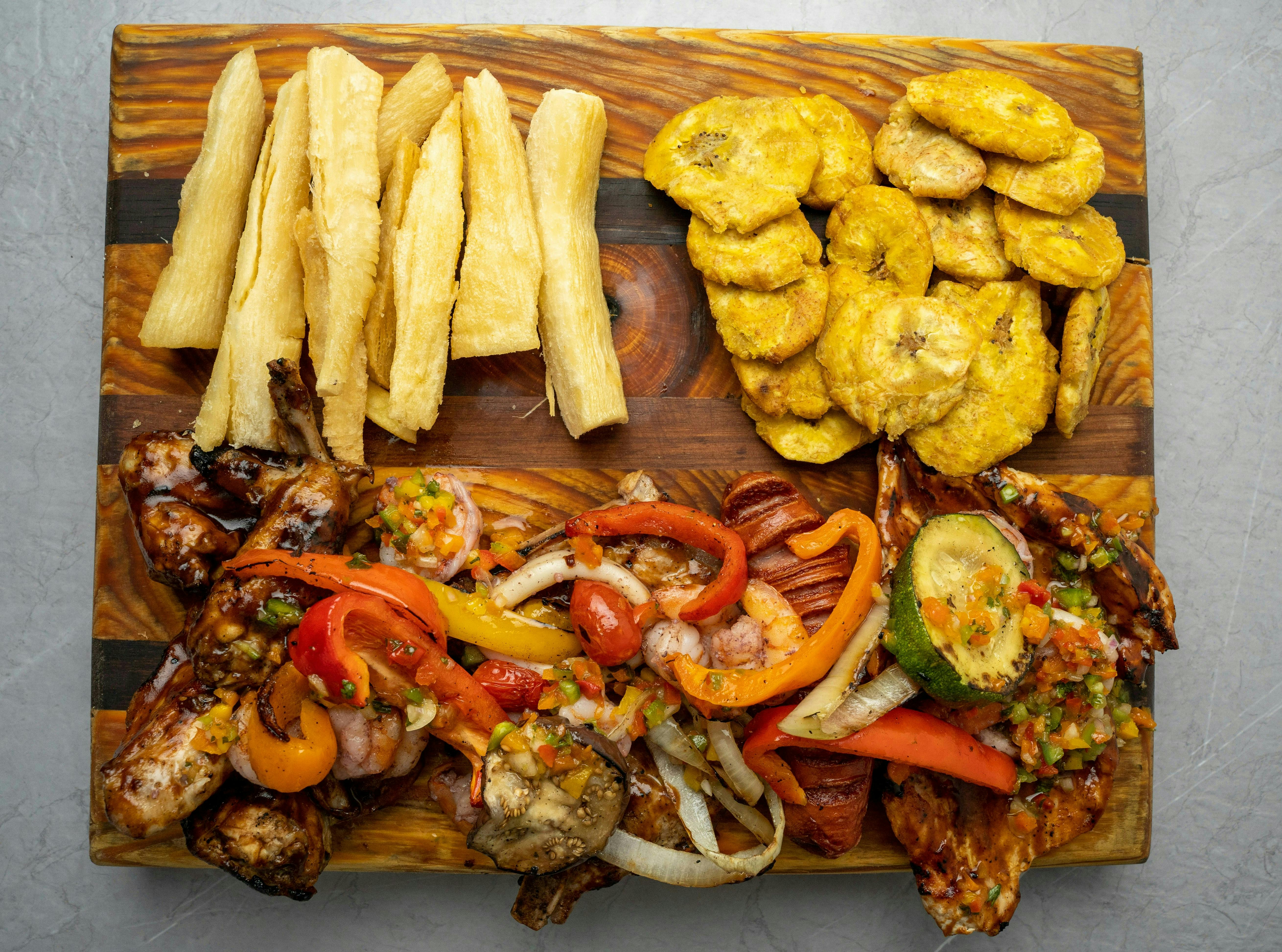 An overhead shot of a large wooden platter filled with various foods, including grilled meat, shrimp, and vegetables, along with fried plantains and what appear to be fried yuca sticks.