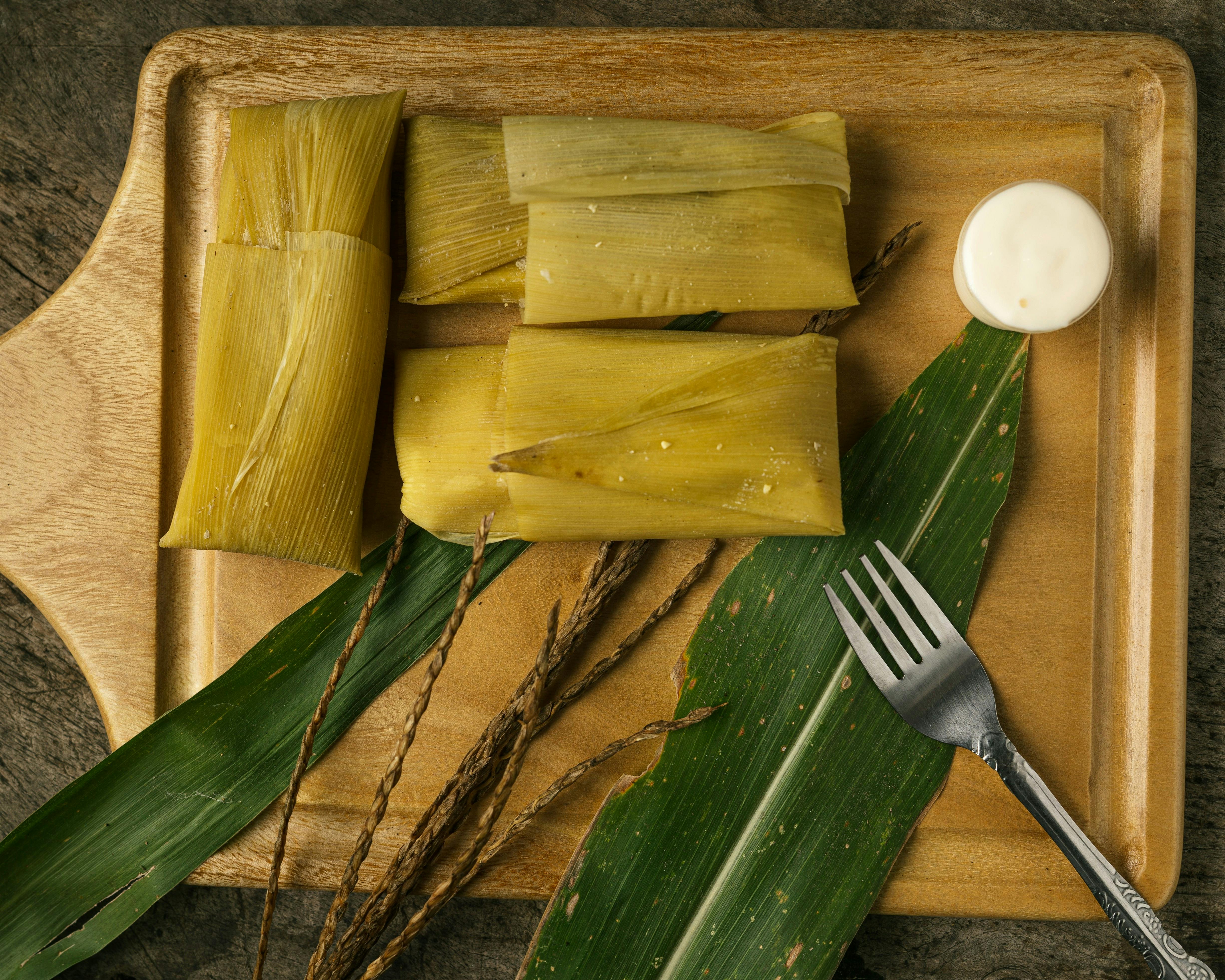 A full-frame, overhead shot of three large corn husks, some with corn silk still attached, lying on a wooden cutting board with a small, round dish of white cheese and a fork.