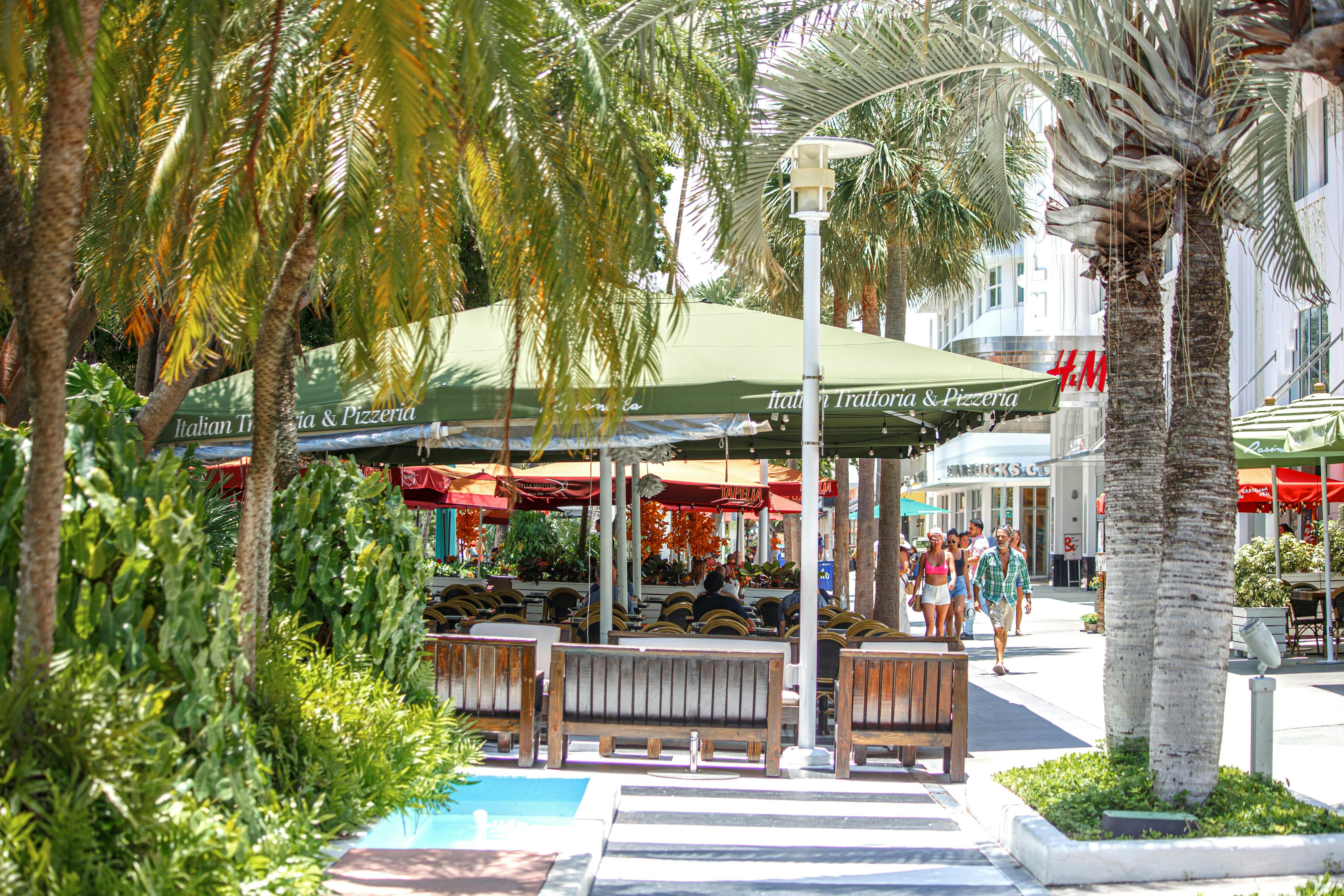 An outdoor cafe with a large green umbrella and tables with red umbrellas. Palm trees line a brick walkway, and people are visible in the background.