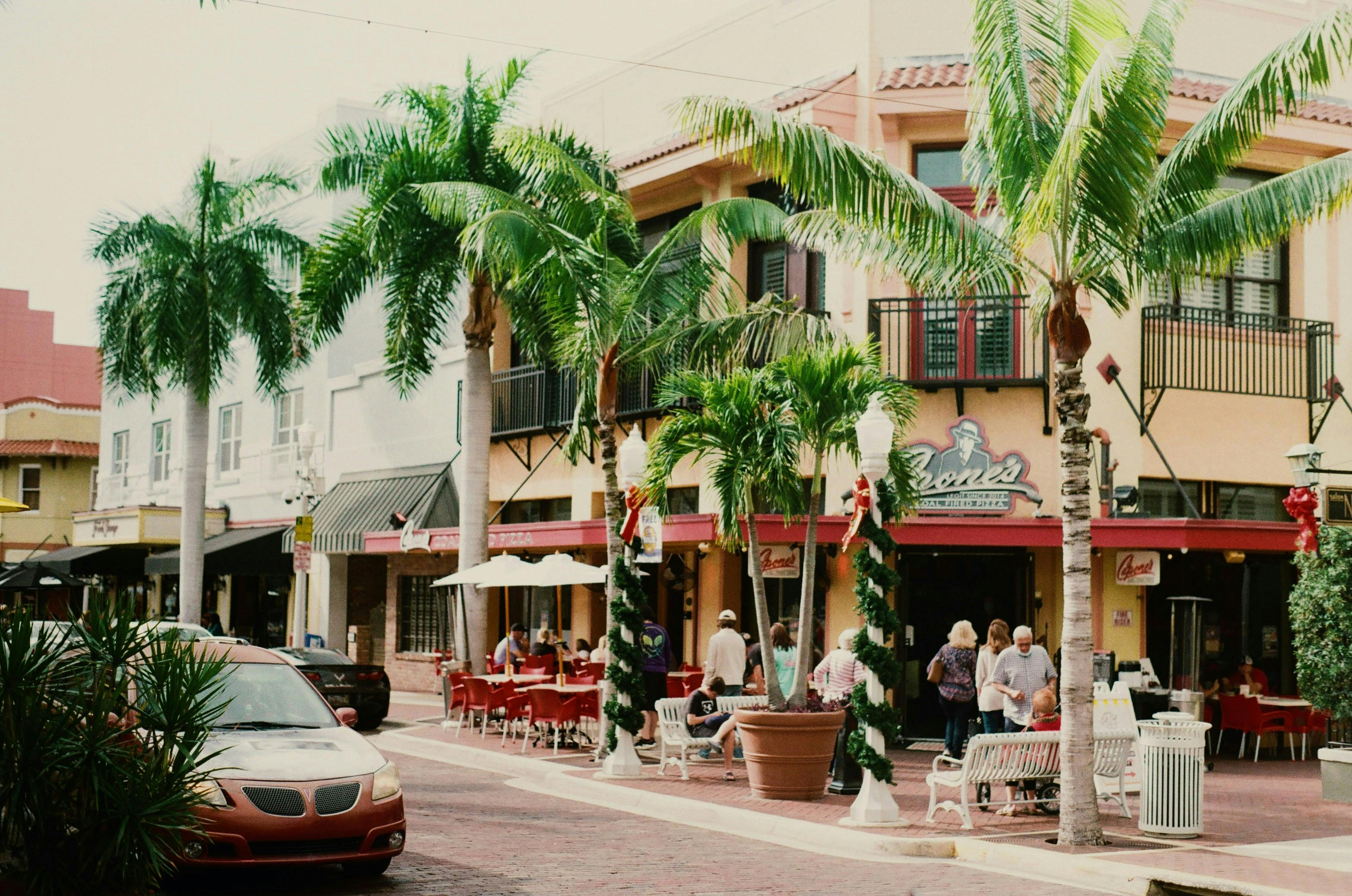 A long shot of a street lined with shops, restaurants, and palm trees. People are sitting at outdoor tables on the sidewalk.