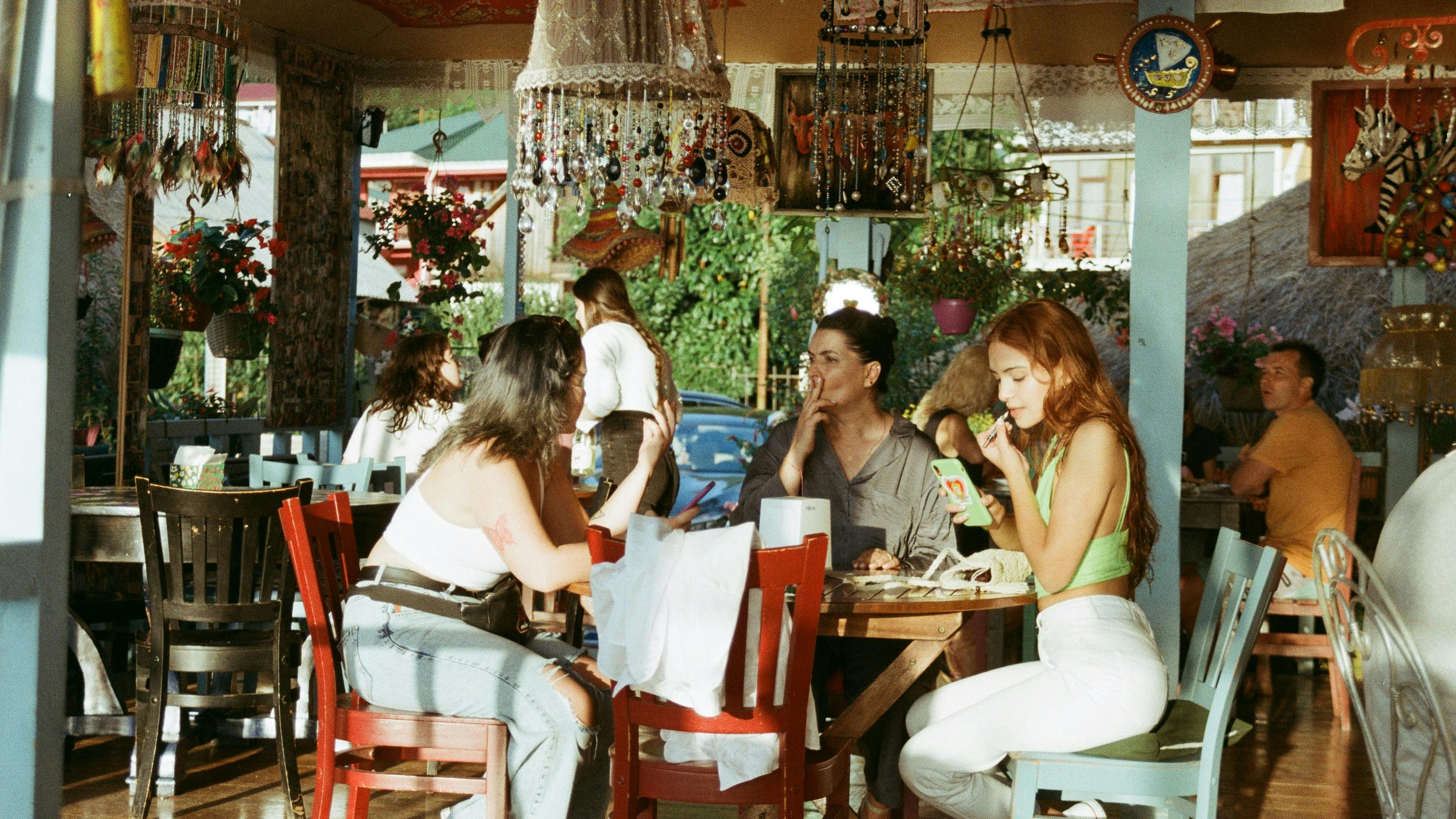 A group of friends is sitting at a wooden table in a colorfully decorated cafe or restaurant with chandeliers and other ornate decorations hanging from the ceiling.