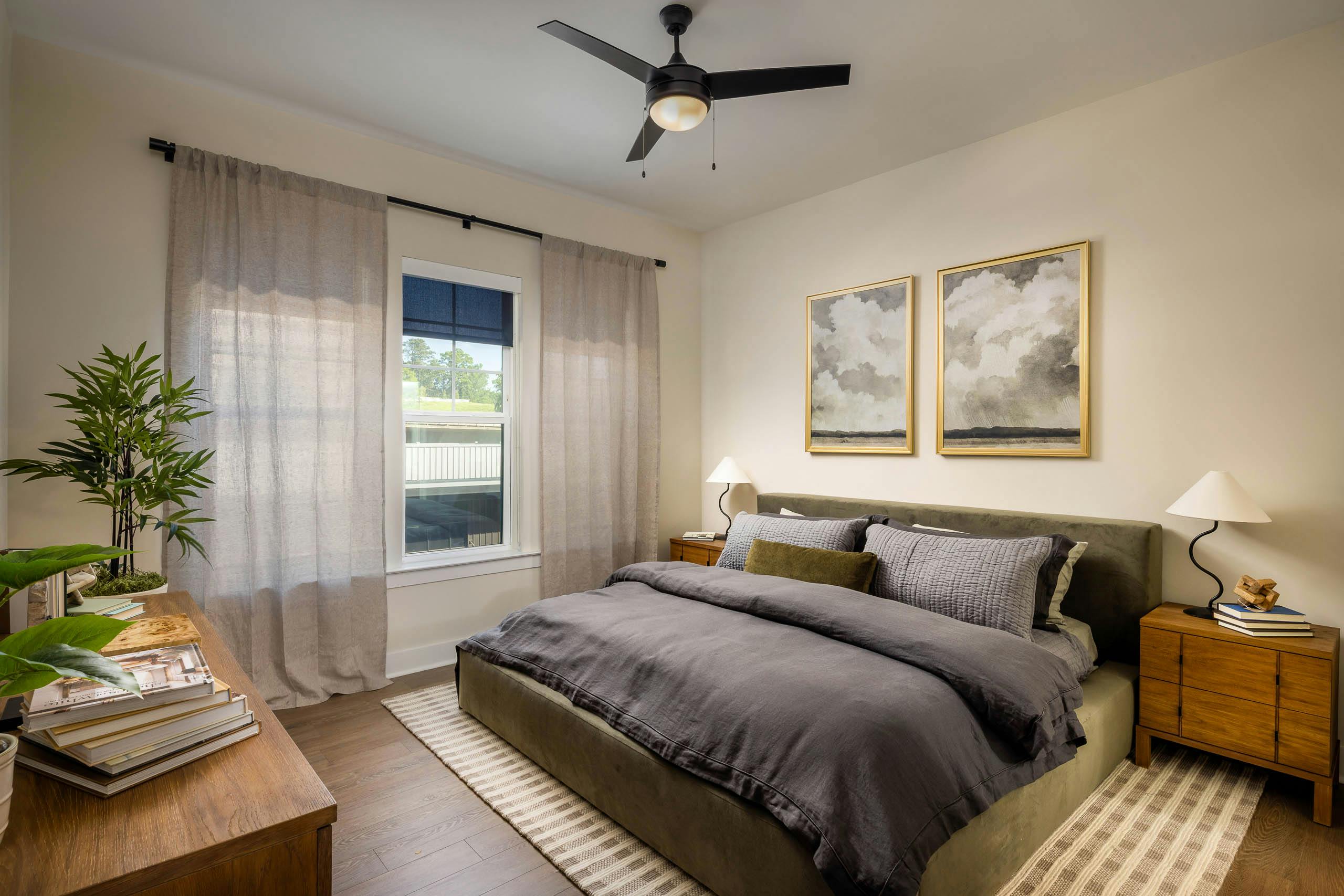 Bedroom at AMLI Brookhaven apartments with hardwood flooring and a ceiling fan and a bed with a grey comforter blanket and a window.