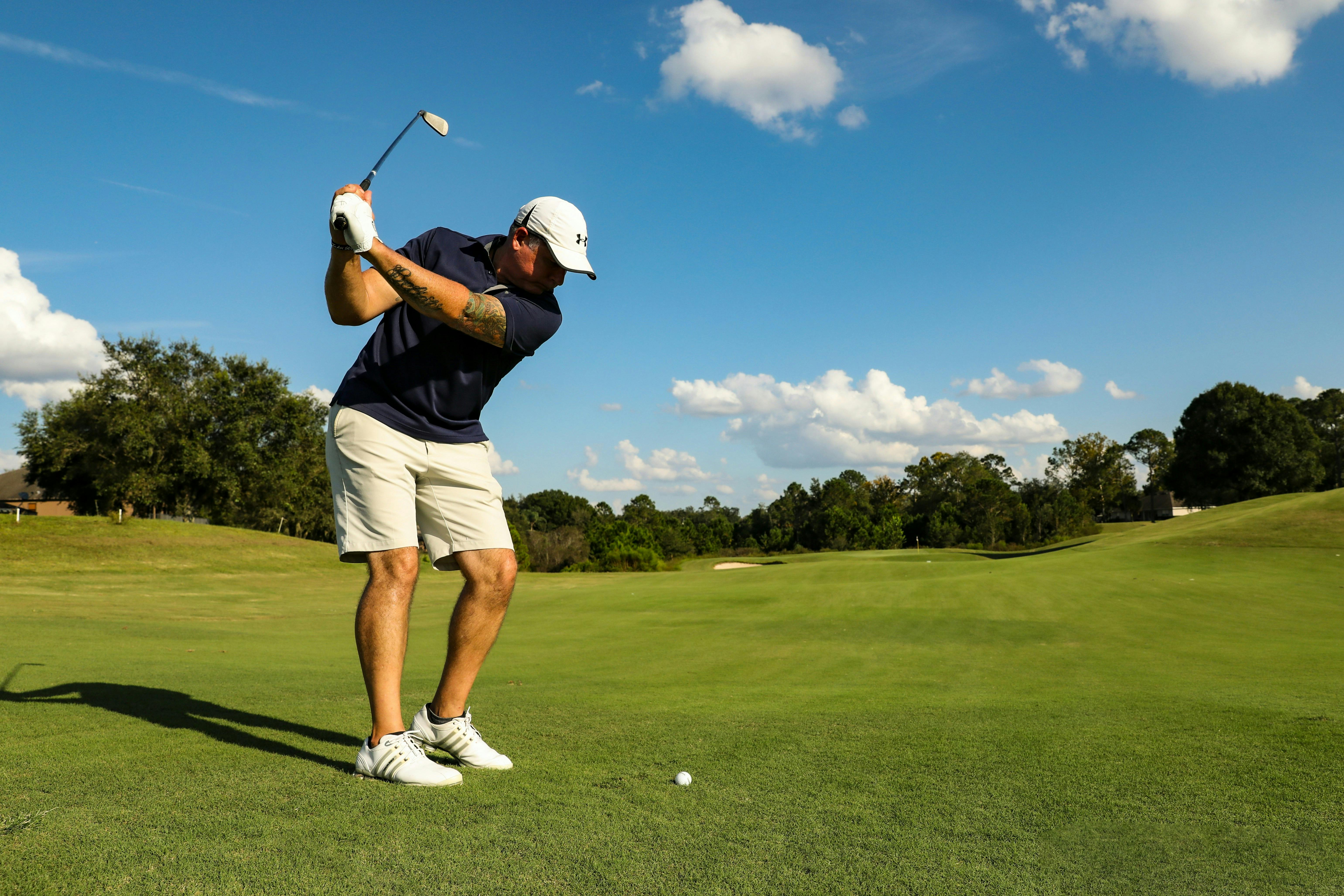 A golfer in a white hat and navy shirt is mid-swing on a green golf course under a clear blue sky.