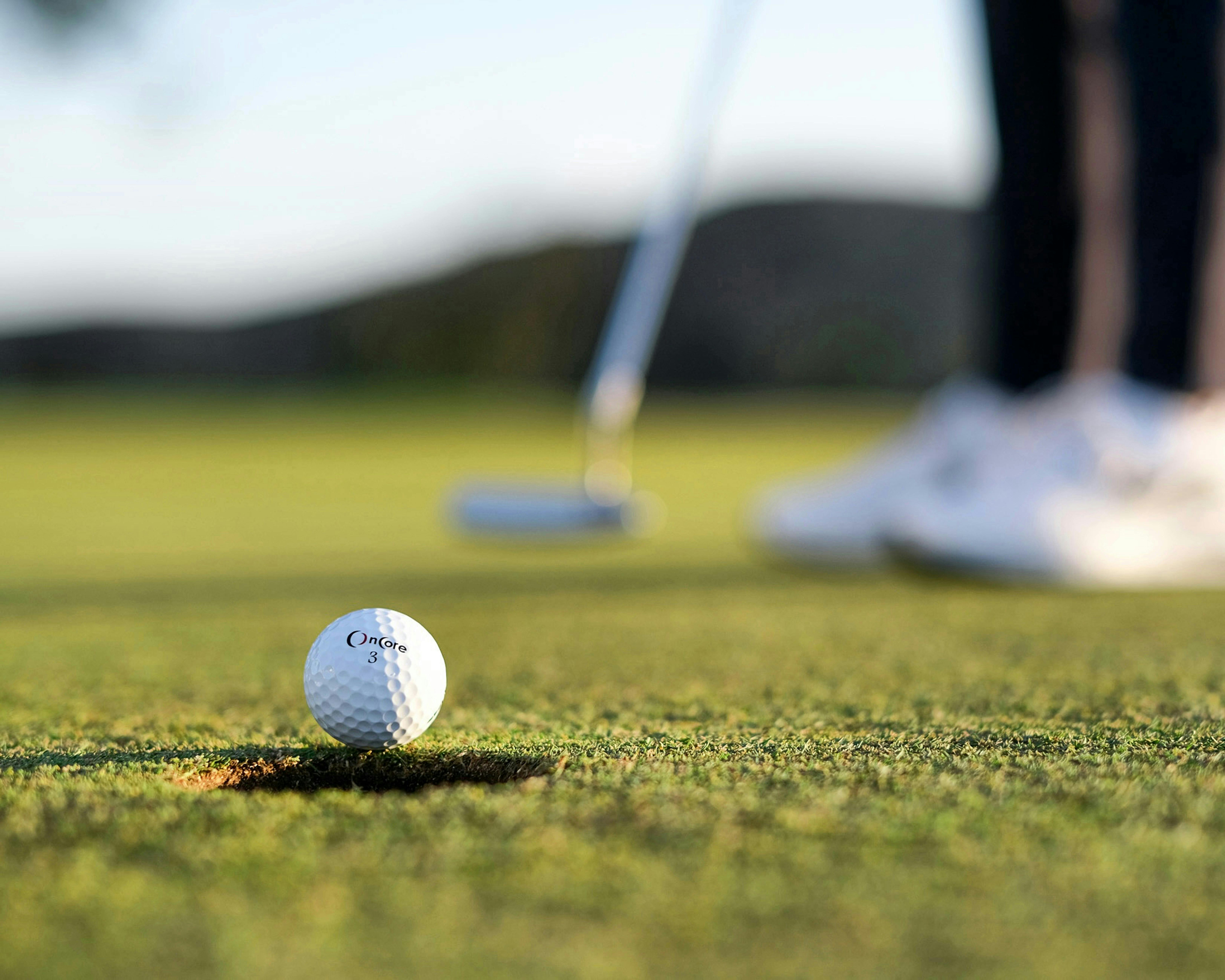 A close-up, low-angle shot of a golf ball with the brand name "Oncore" on it, sitting at the edge of a hole on a green. A golfer's feet and putter are blurred in the background.