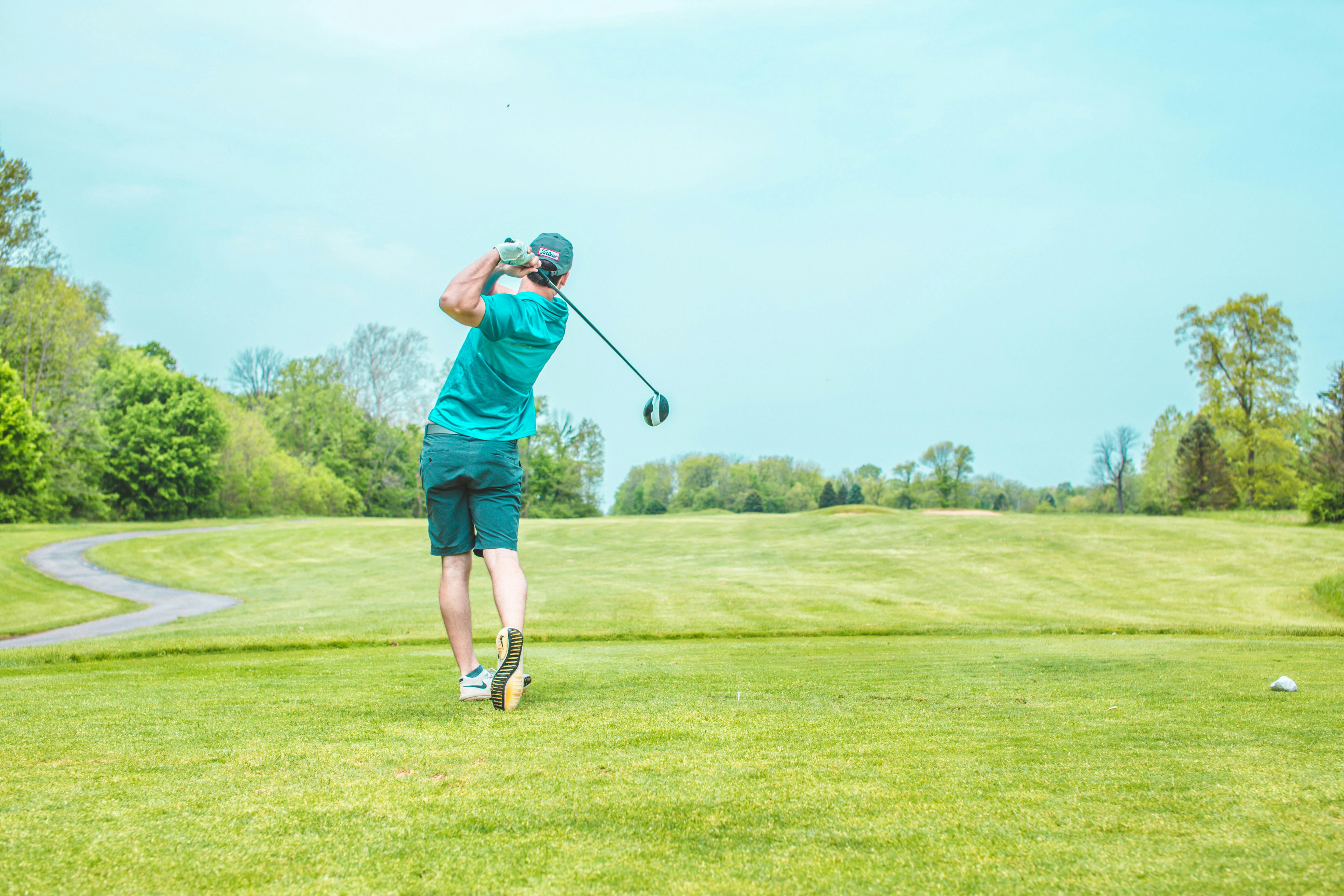 A golfer in a blue shirt and dark shorts is mid-swing with a driver on a wide green fairway under a bright sky.