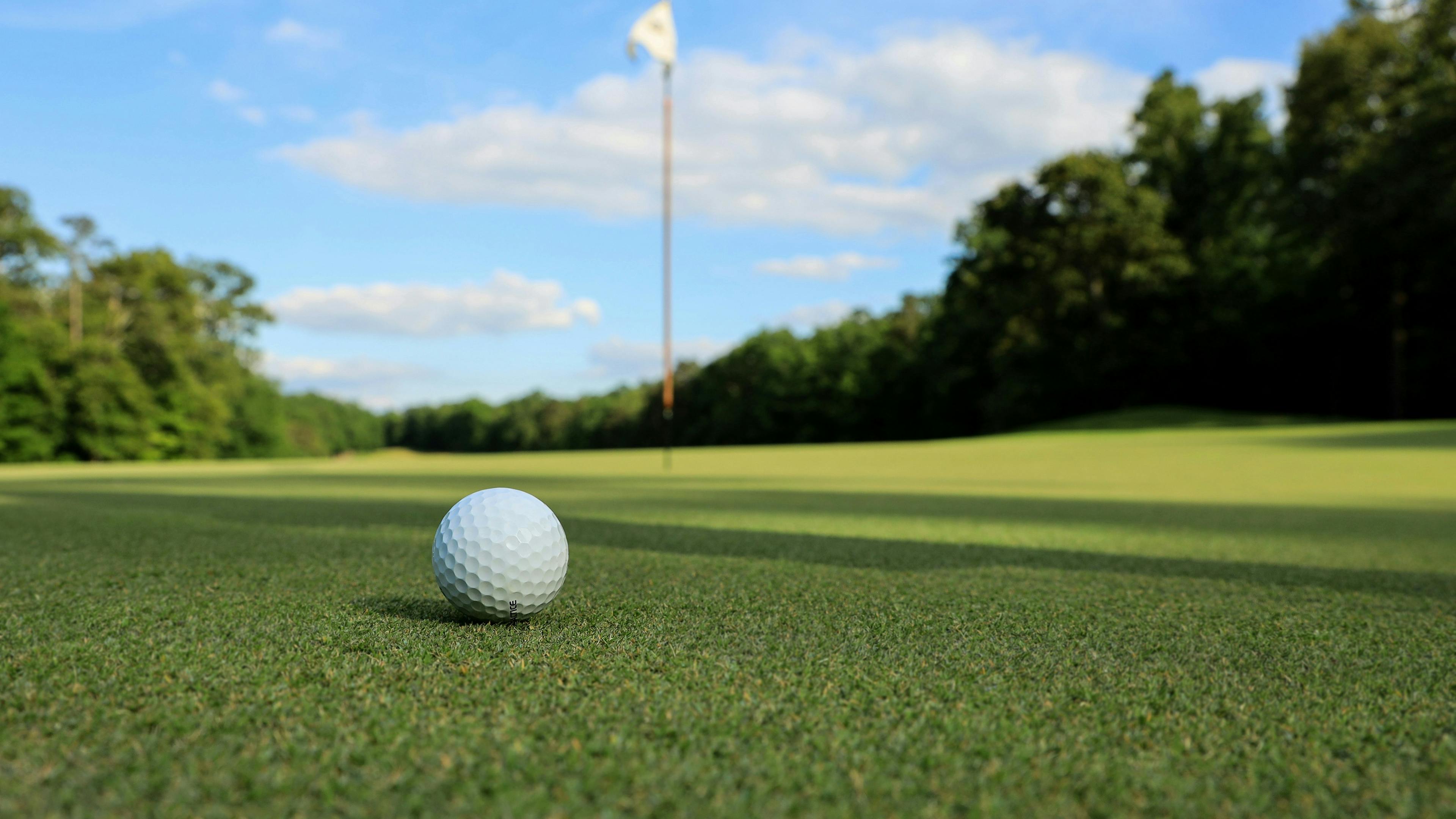 A low-angle shot of a golf ball on a green, with a flagstick in the distance. The grass is lush and green under a bright blue sky with scattered clouds.
