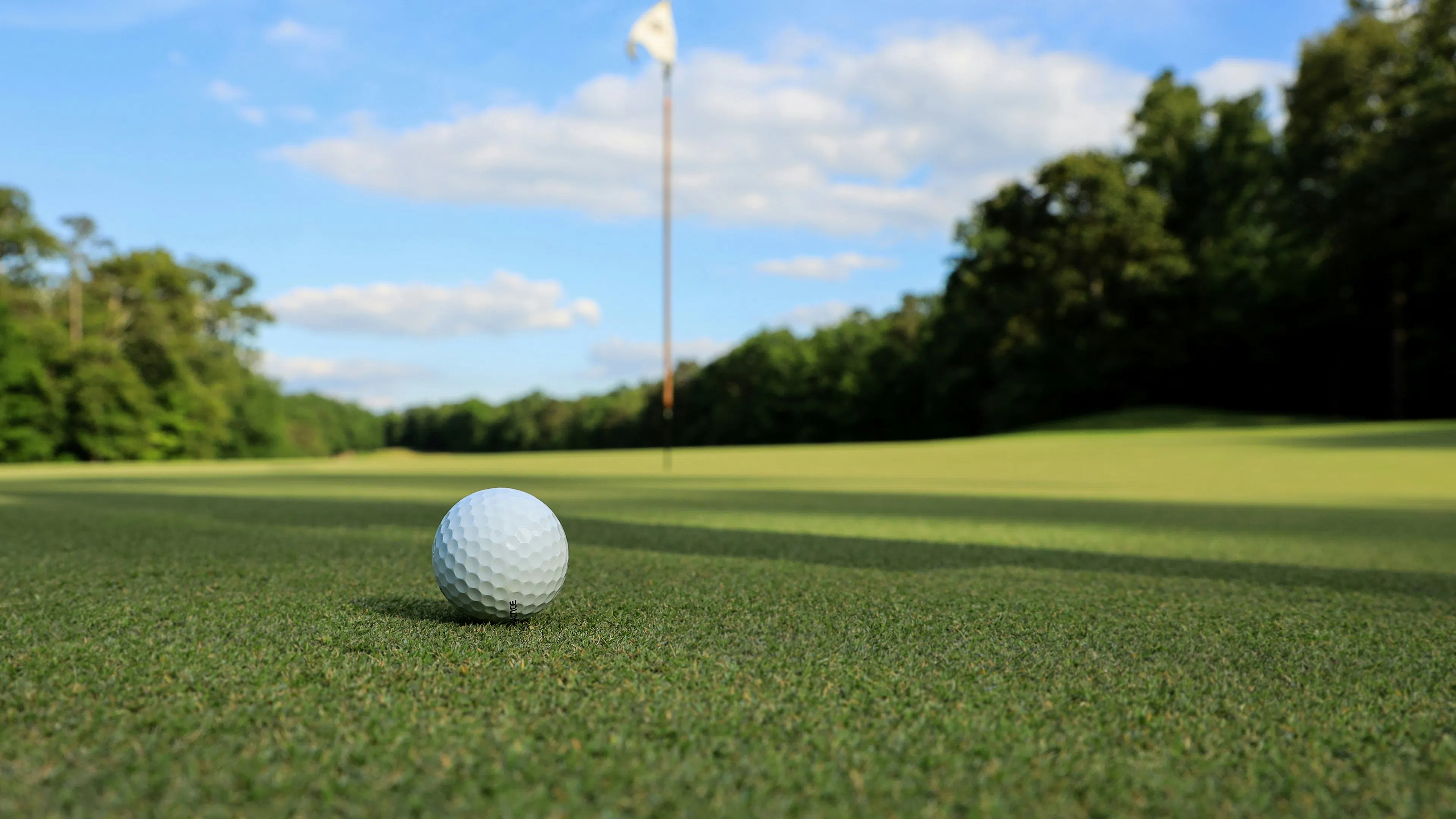 A low-angle shot of a golf ball on a green, with a flagstick in the distance. The grass is lush and green under a bright blue sky with scattered clouds.