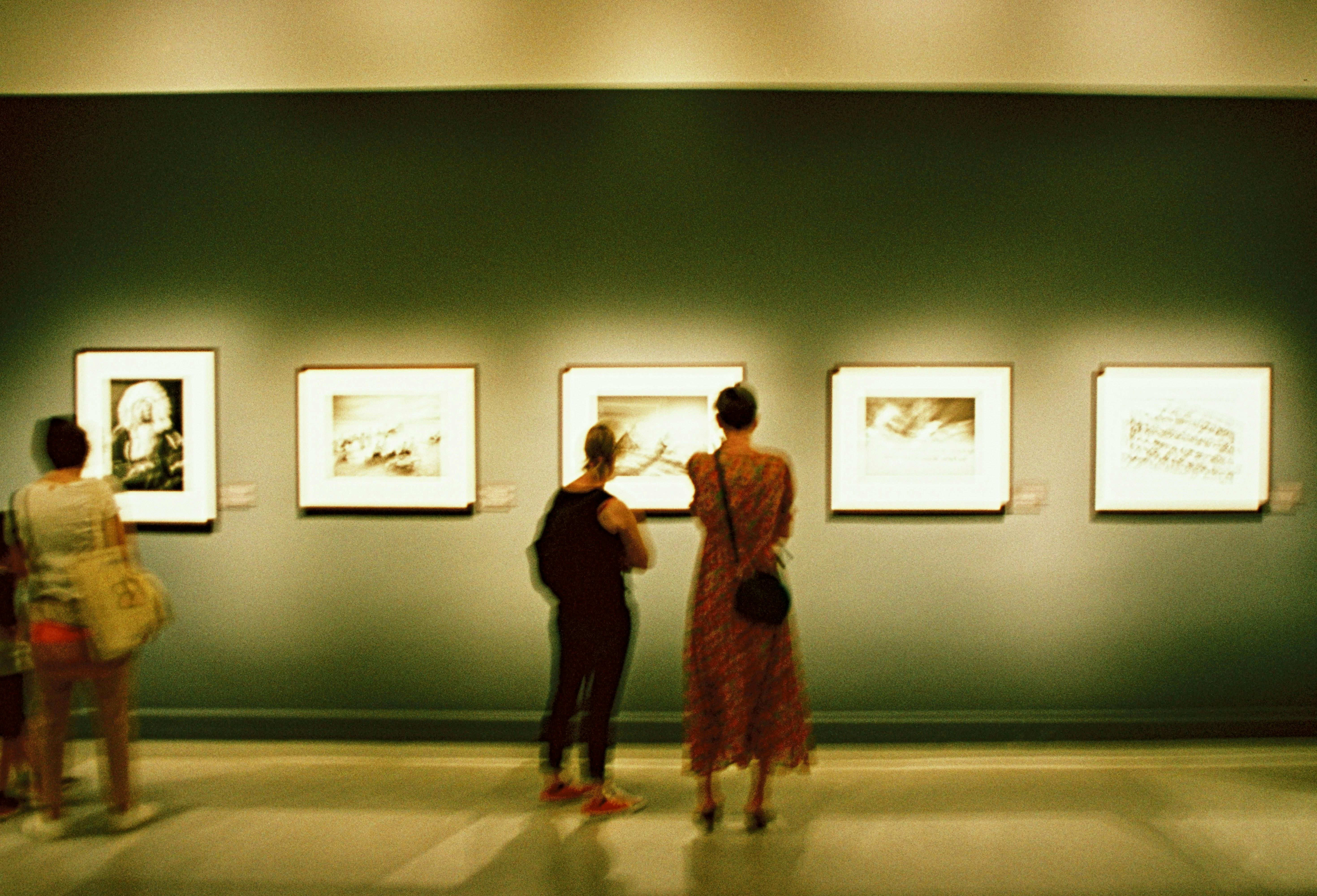 A blurry, grainy shot of people in an art gallery, viewed from behind, looking at a series of five framed black and white photographs.