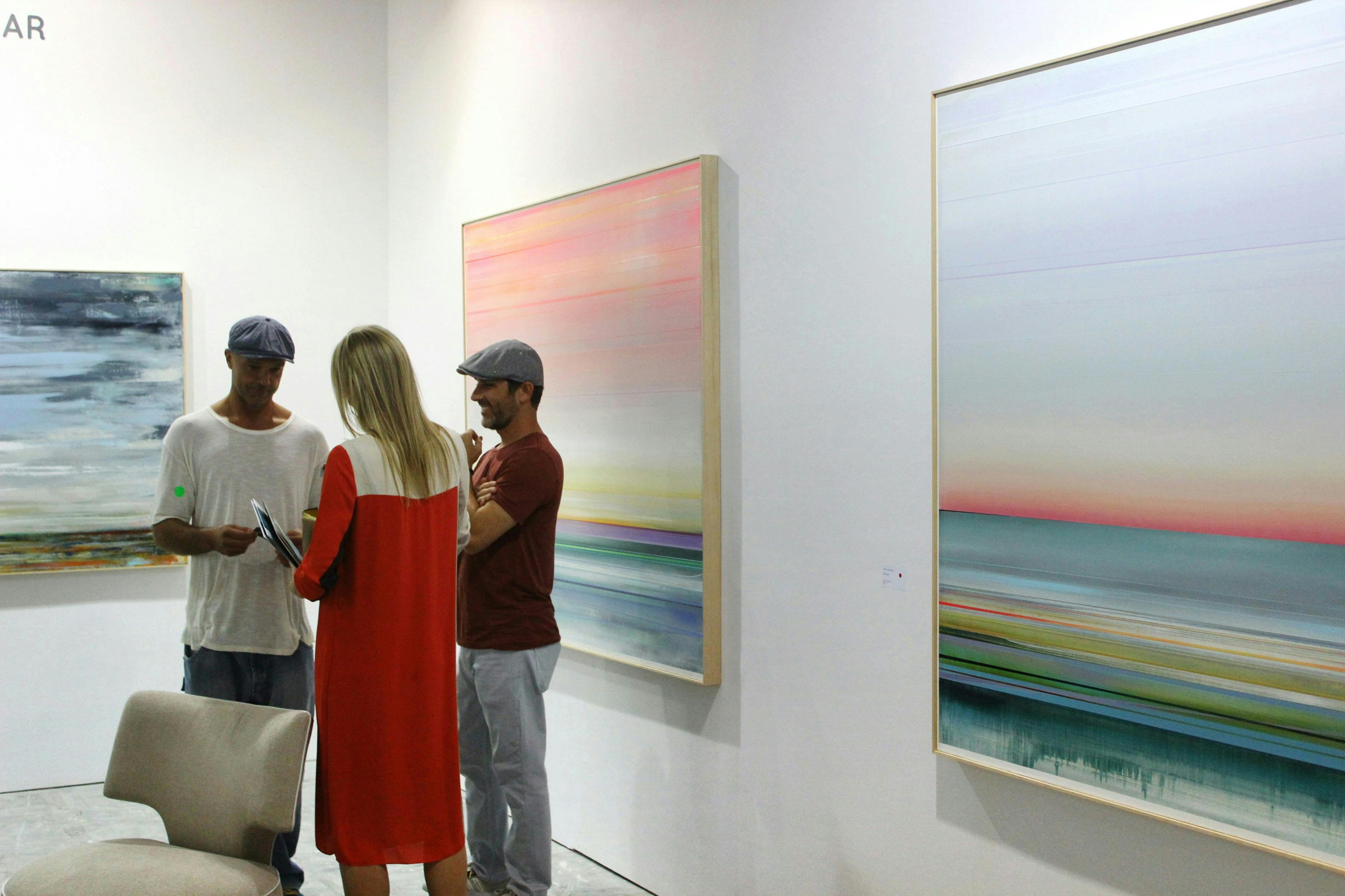 Three people are standing and talking in a modern art gallery with white walls. They are standing near two large, abstract paintings that feature horizontal lines and a blend of vibrant colors.