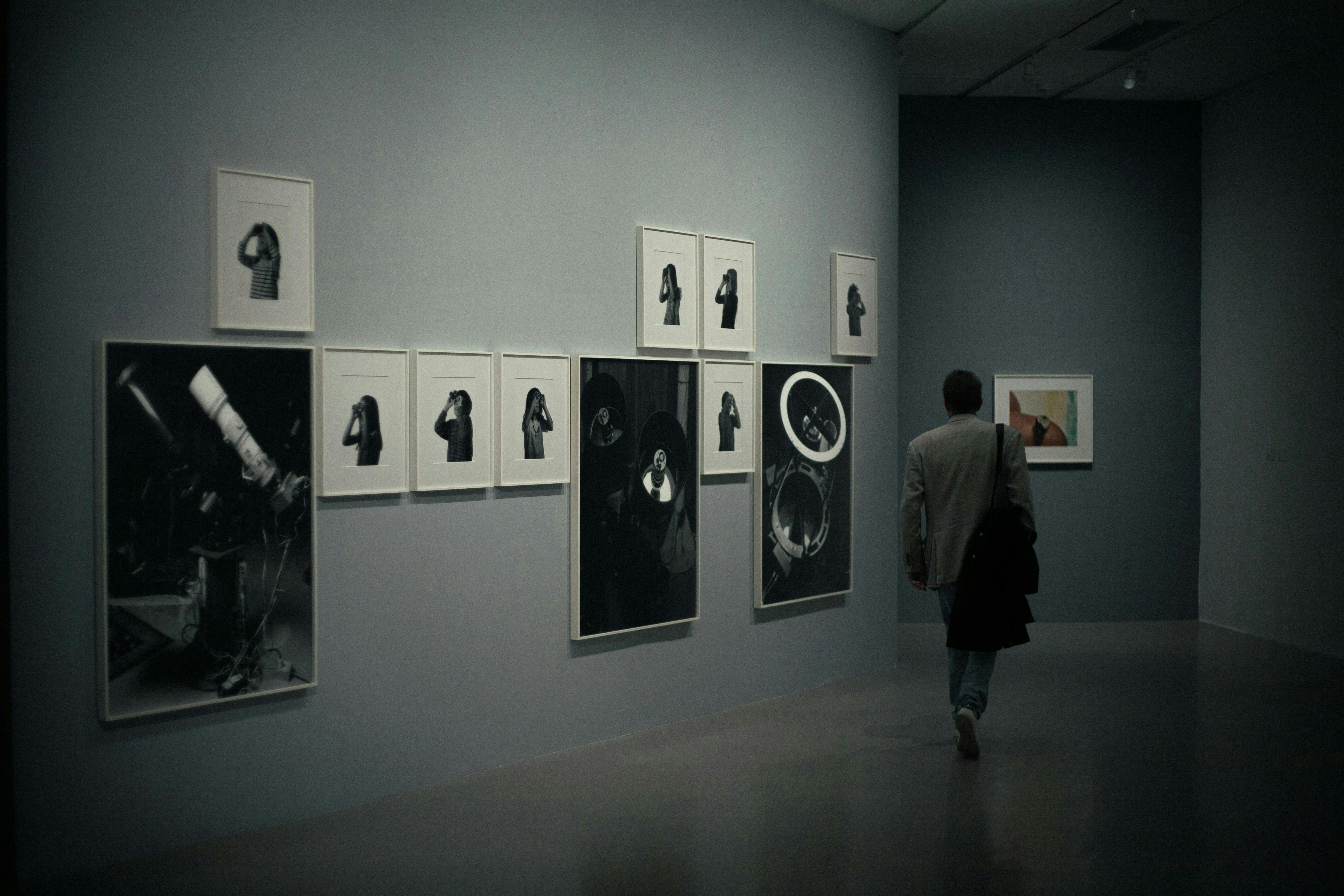 A man with a satchel is walking through a dimly lit art gallery with dark gray walls, looking at a collection of black and white framed prints of various sizes.