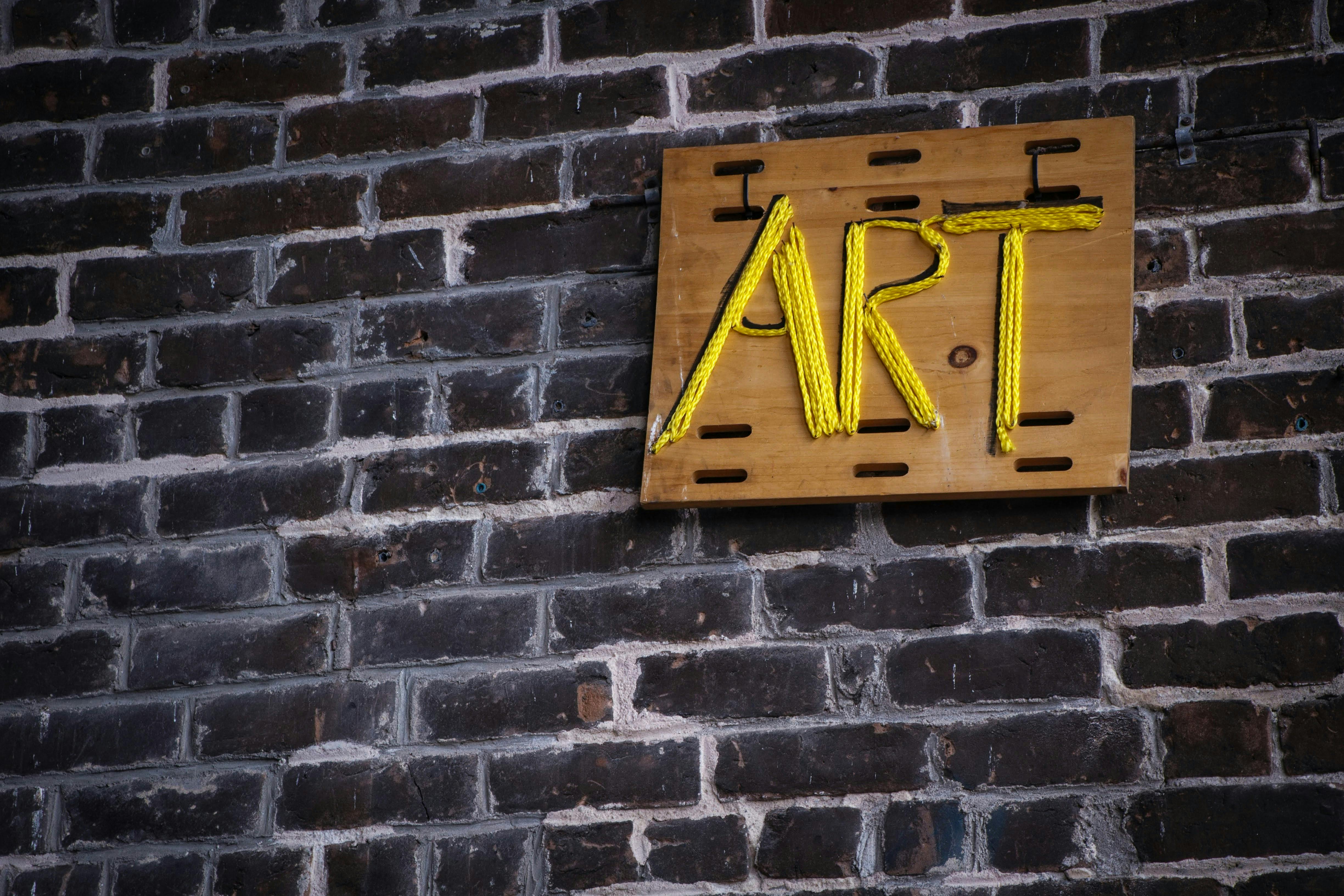 A sign with the word "ART" spelled out in yellow rope on a wooden board is mounted on a dark brick wall.