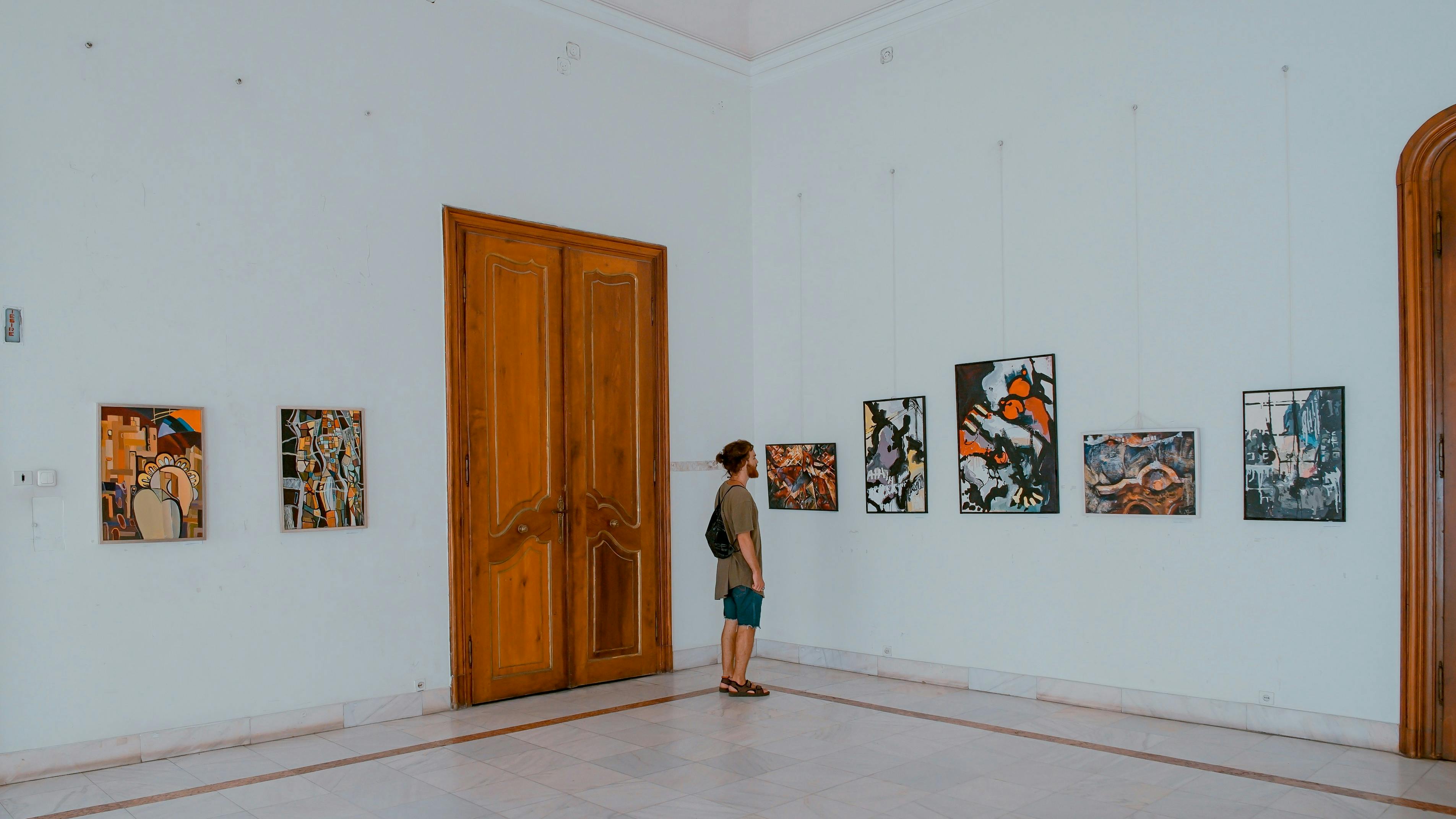 A person stands in a large, white-walled room with a high ceiling and a marble floor, looking at a row of colorful framed paintings on the wall. A tall, carved wooden double door is visible on the left.