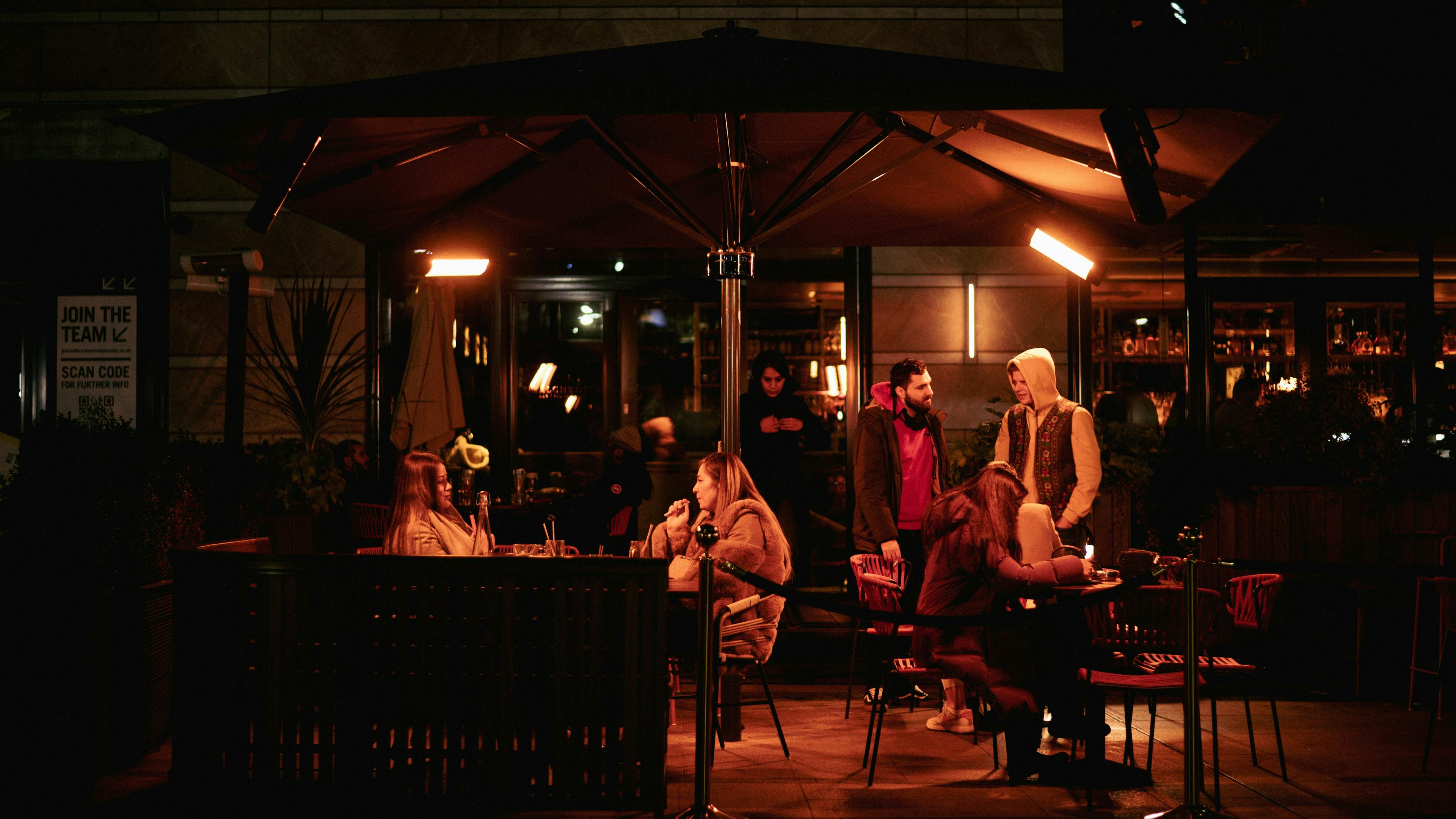 A dimly lit outdoor patio of a restaurant at night. Groups of people are seated at tables under a large umbrella, with a few others standing nearby. Heat lamps provide a warm glow.