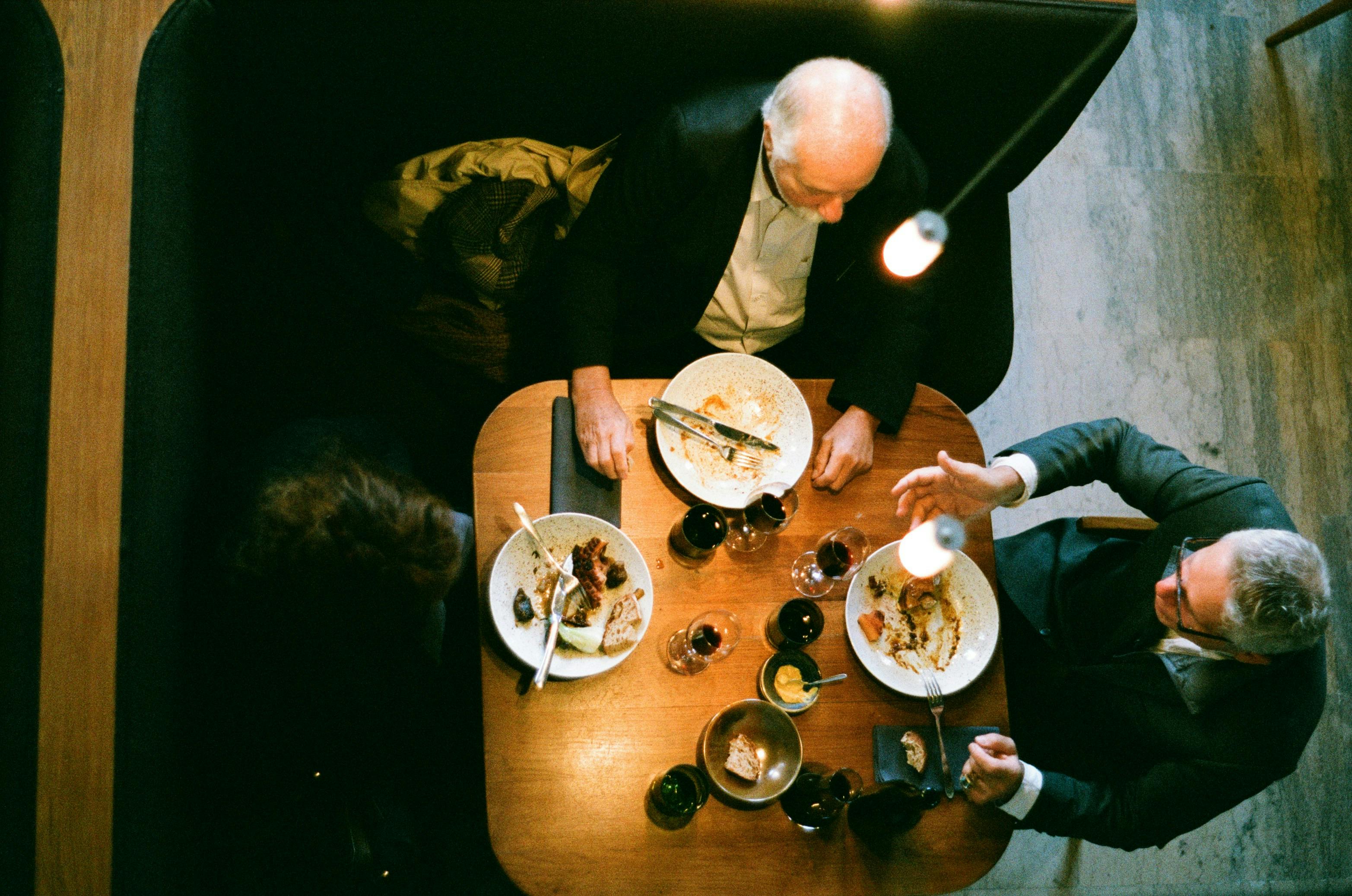 An overhead shot of two men in suits eating at a booth in a restaurant. Their plates are mostly empty, and there are several wine glasses on the table.