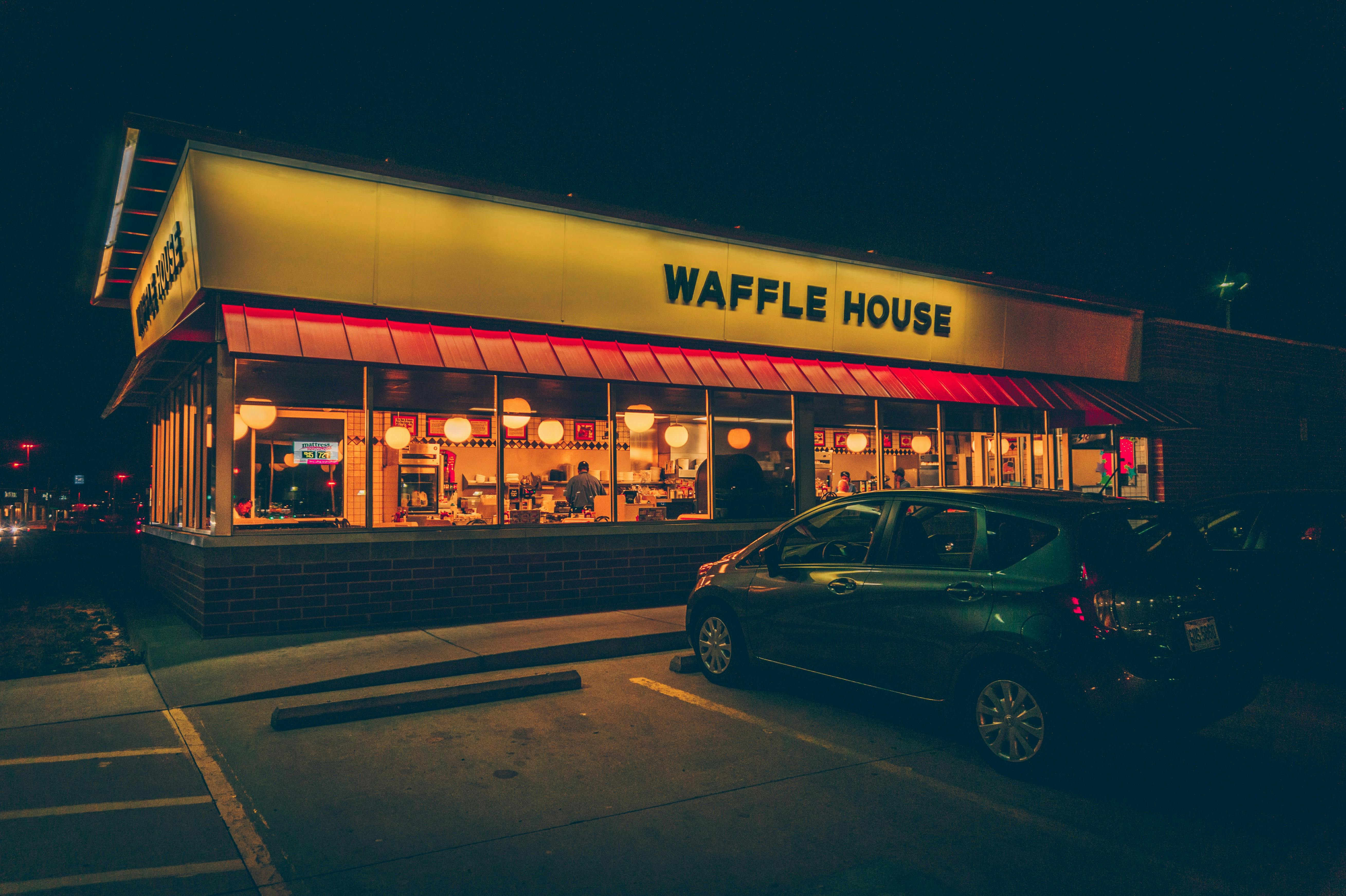 A low-angle, street-level shot of a Waffle House restaurant at night. The building is brightly lit with warm lights, and a car is parked in the foreground. The sign "WAFFLE HOUSE" is prominently displayed.