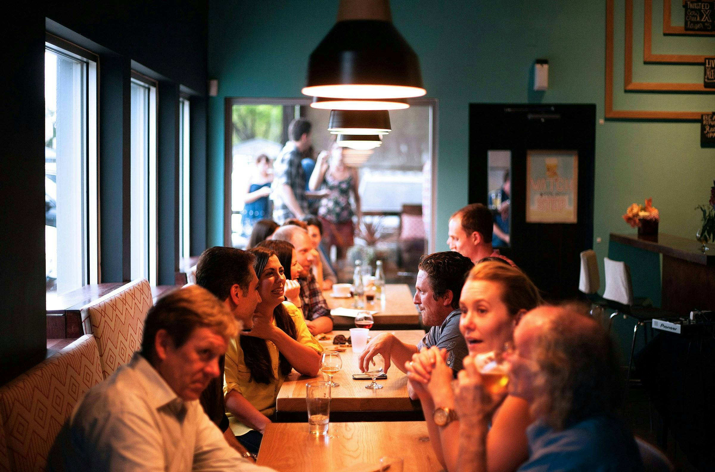 A long-shot of a group of people sitting at a long table in a restaurant. They are talking and laughing while eating and drinking wine.