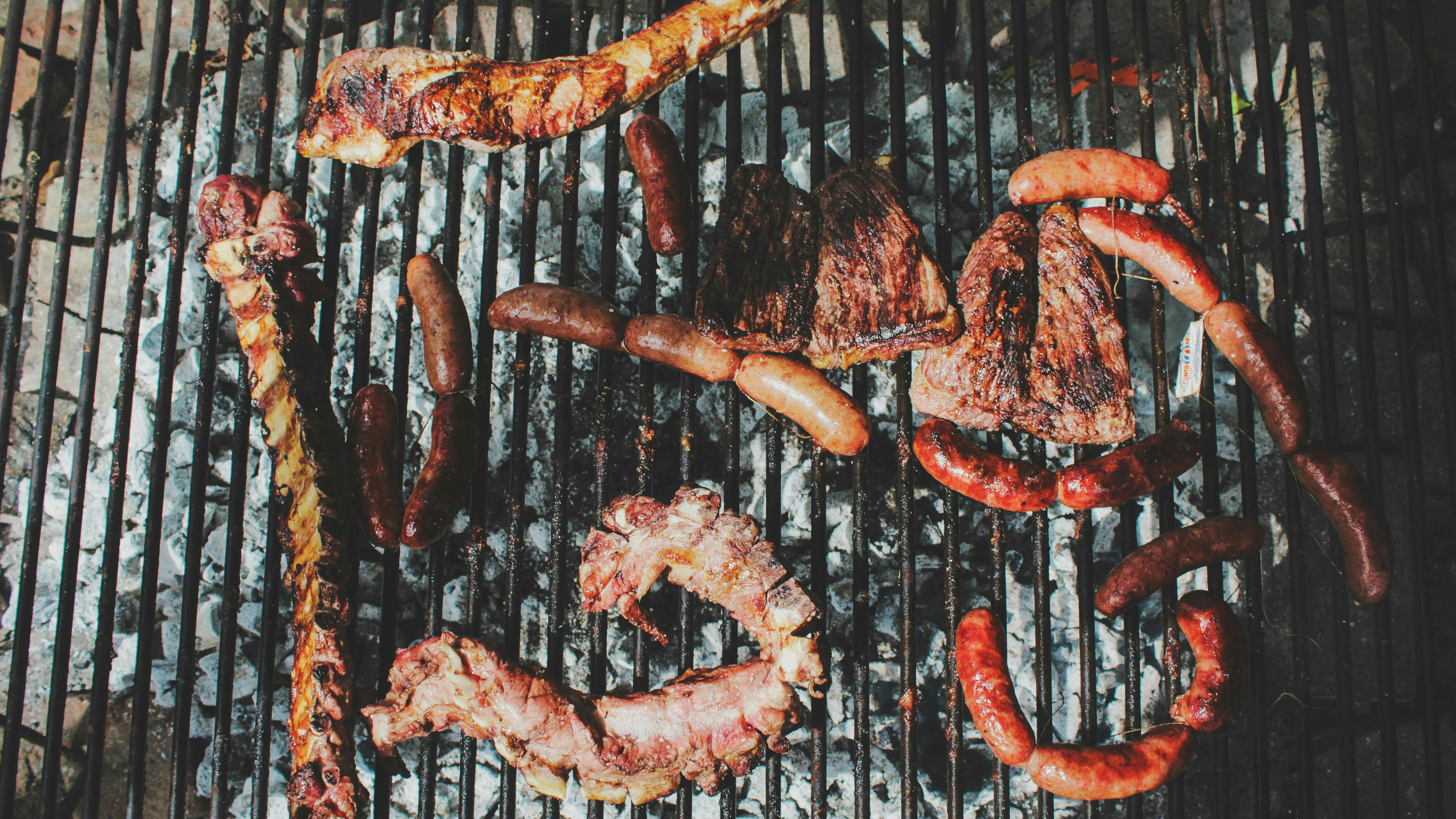 A full-frame, overhead shot of various cuts of meat, including ribs and sausages, cooking on a wire grill over hot coals.