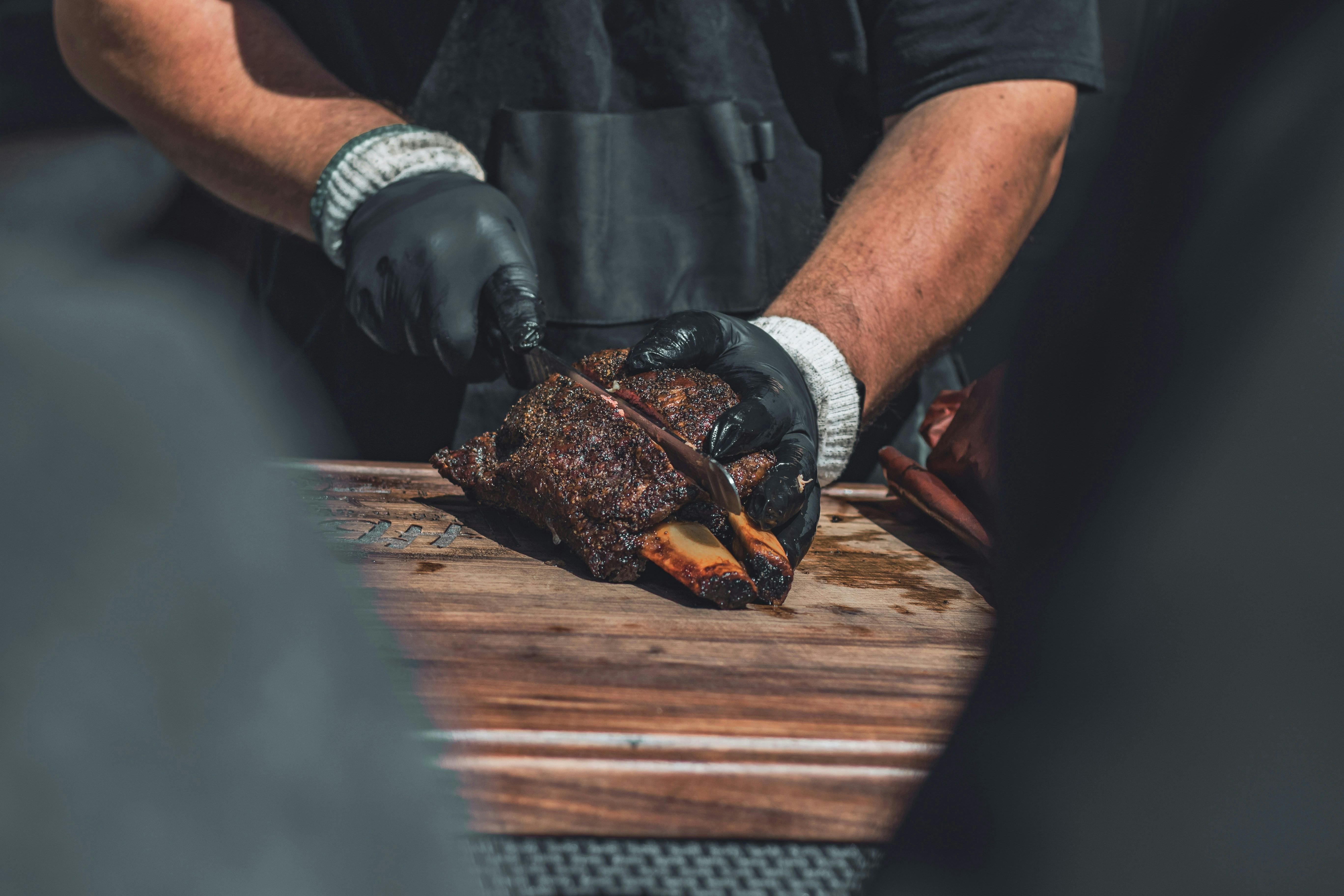 A close-up, high-angle shot of a person wearing black gloves slicing a large, meaty bone-in rib on a wooden cutting board with a knife.