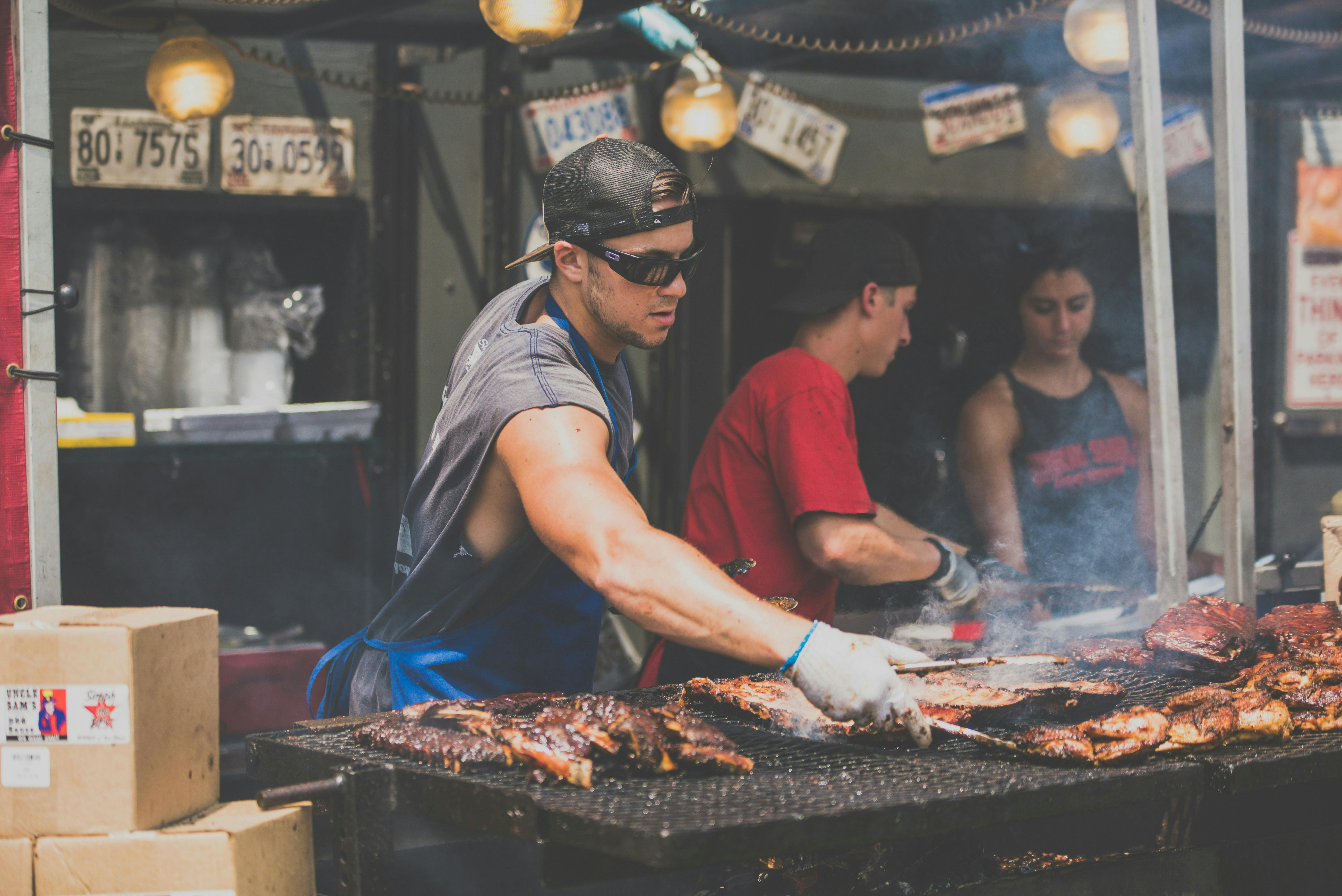 A man in a blue apron and sunglasses is grilling large slabs of meat and ribs on a wide, smoky outdoor grill. Two other people are visible behind him.